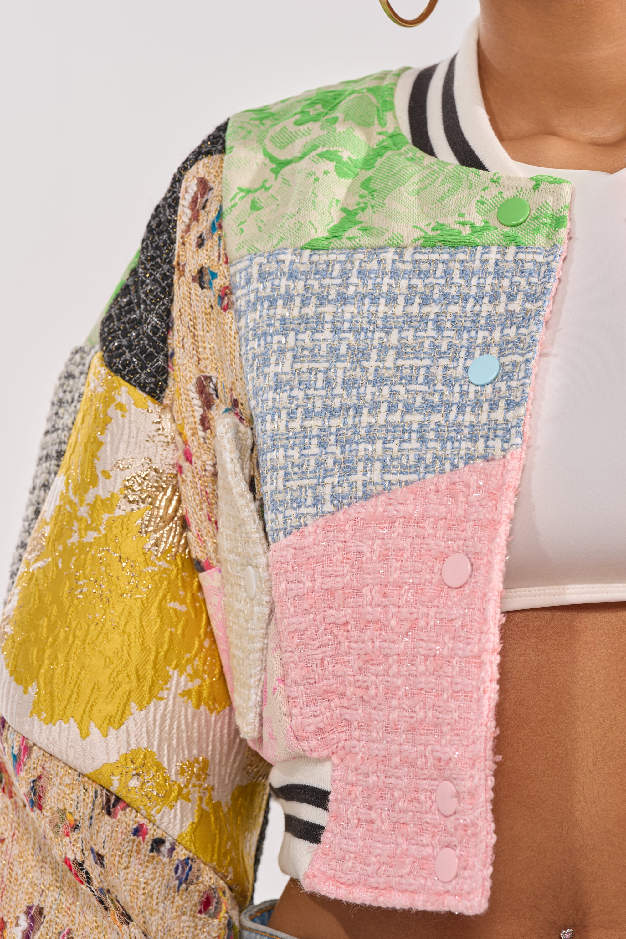 Close-up of a person wearing the ESTELLE PATCHWORK BOMBER, featuring textured pink, blue, yellow, and green fabrics over a white crop top. Striped collar and pink buttons add standout detail to this vibrant statement streetwear piece. 10