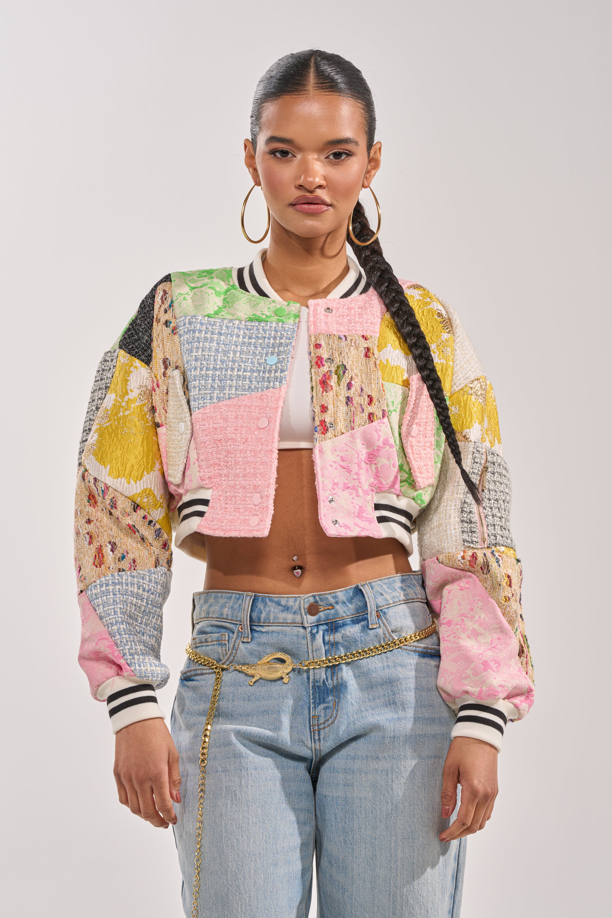 A woman with long braided hair wears the ESTELLE PATCHWORK BOMBER over a white crop top, light blue jeans, a gold chain belt, and large hoop earrings, standing against a plain light background. 1