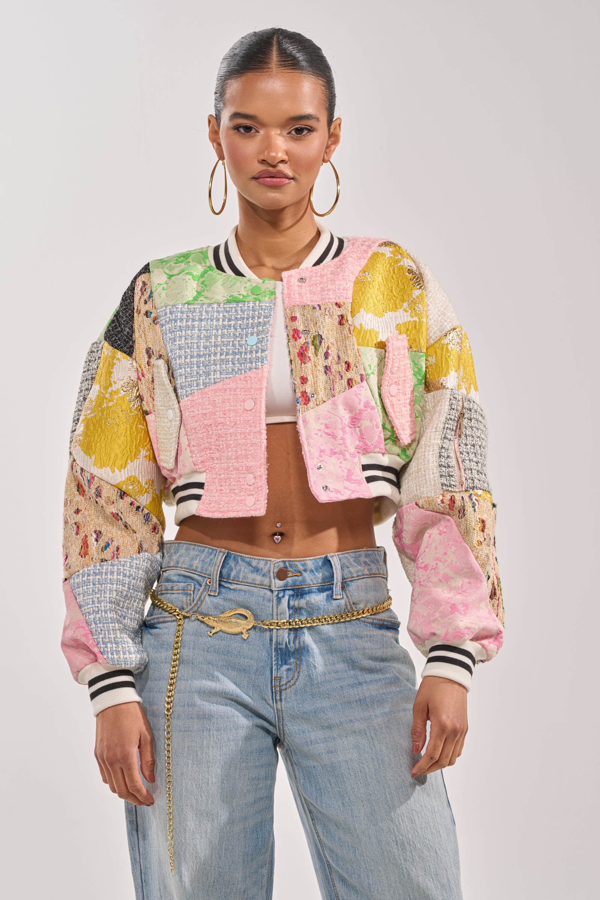 A woman poses against a plain background in the ESTELLE PATCHWORK BOMBER, light blue jeans with a gold chain belt, and large hoop earrings—creating an effortless, statement streetwear look. 2