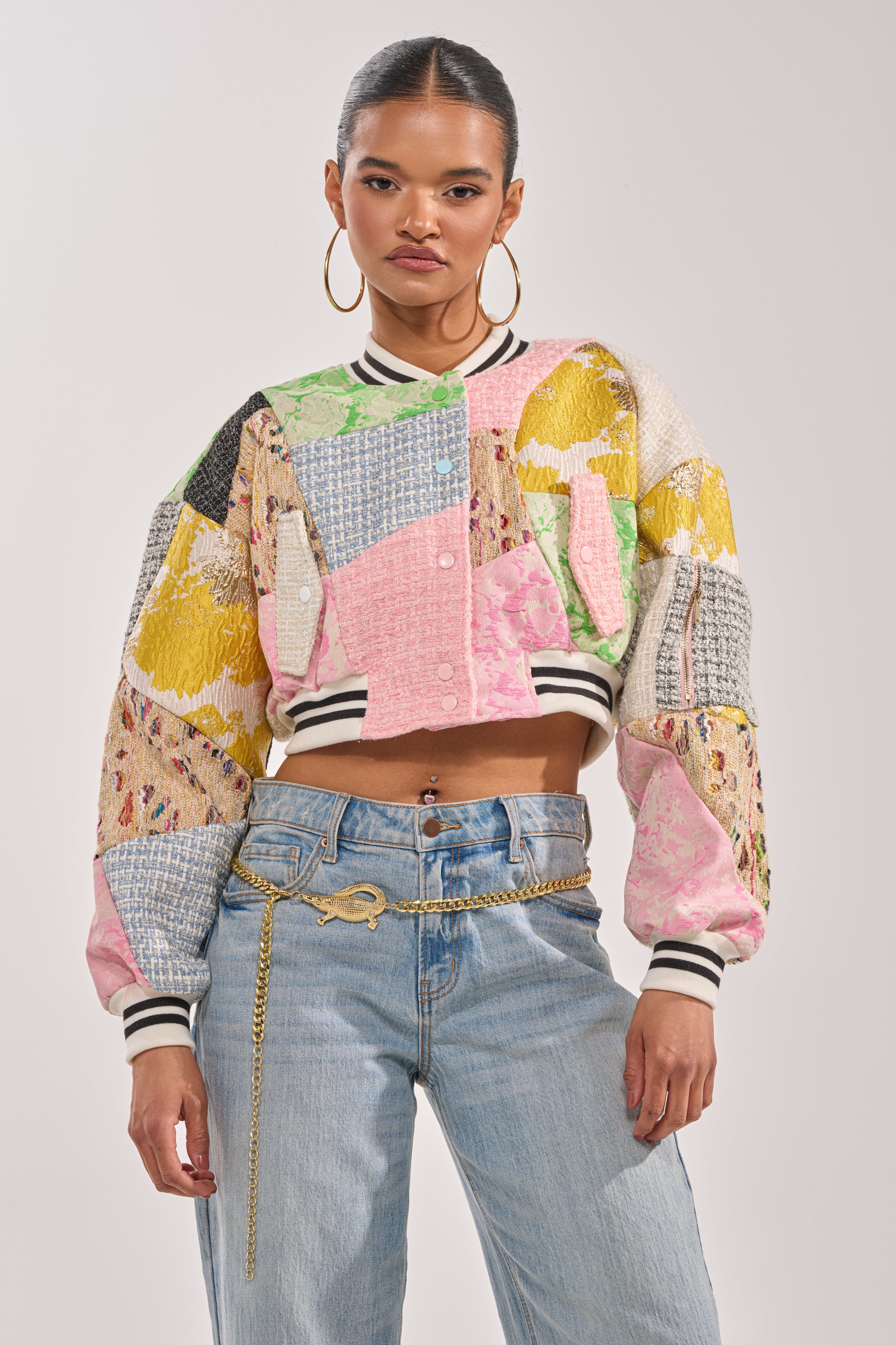 A woman exudes confidence in the ESTELLE PATCHWORK BOMBER, featuring mixed textured fabrics, paired with light-wash high-waisted jeans and a gold chain belt. Her hair is pulled back, completing her bold streetwear look. 3