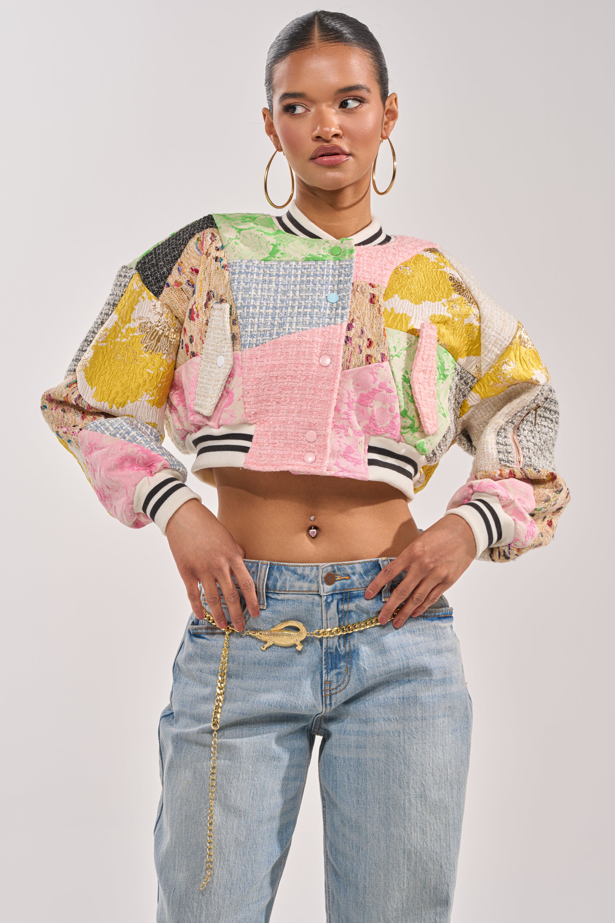 A woman stands confidently in the ESTELLE PATCHWORK BOMBER, styled over light blue jeans with a gold chain belt. Her hair is tied back as she poses with hands on her waist against a plain background. 4