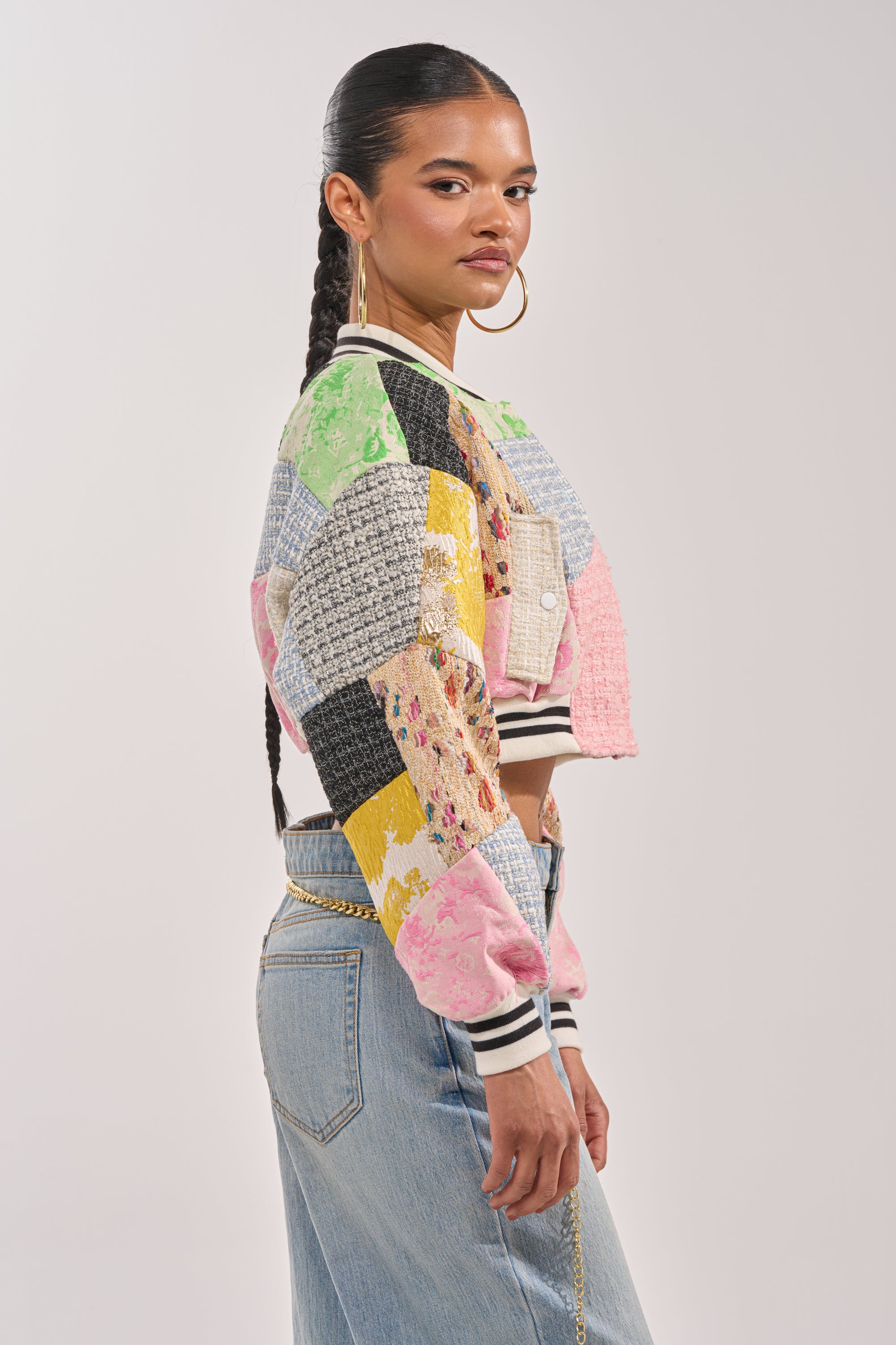 A woman with long braided hair models the ESTELLE PATCHWORK BOMBER and light blue jeans, accessorized with large hoop earrings. She stands sideways against a plain light background, creating a bold statement streetwear look. 5