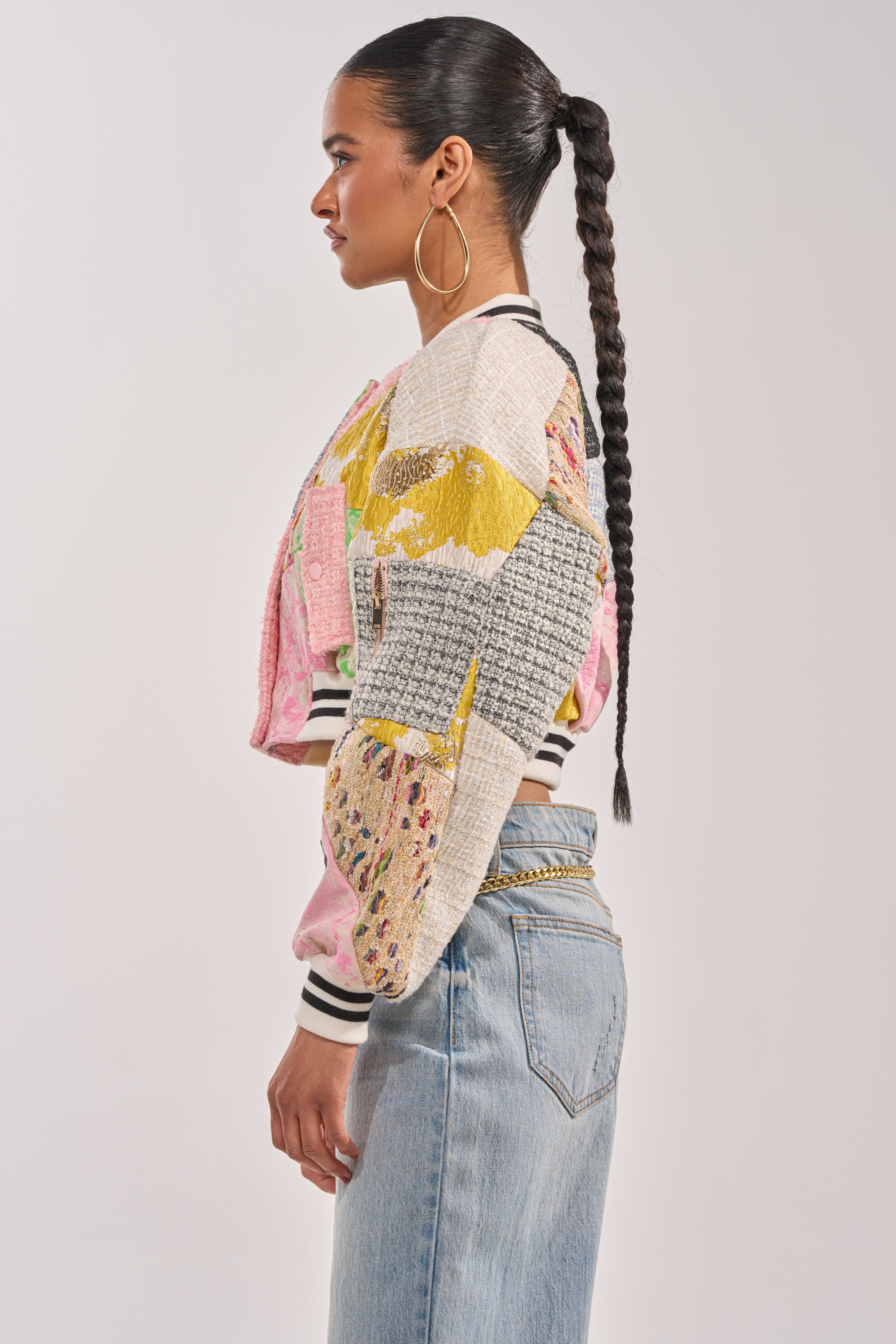 A woman with a long braided ponytail and large hoop earrings models the ESTELLE PATCHWORK BOMBER and light blue jeans, standing in profile against a plain light background. 6