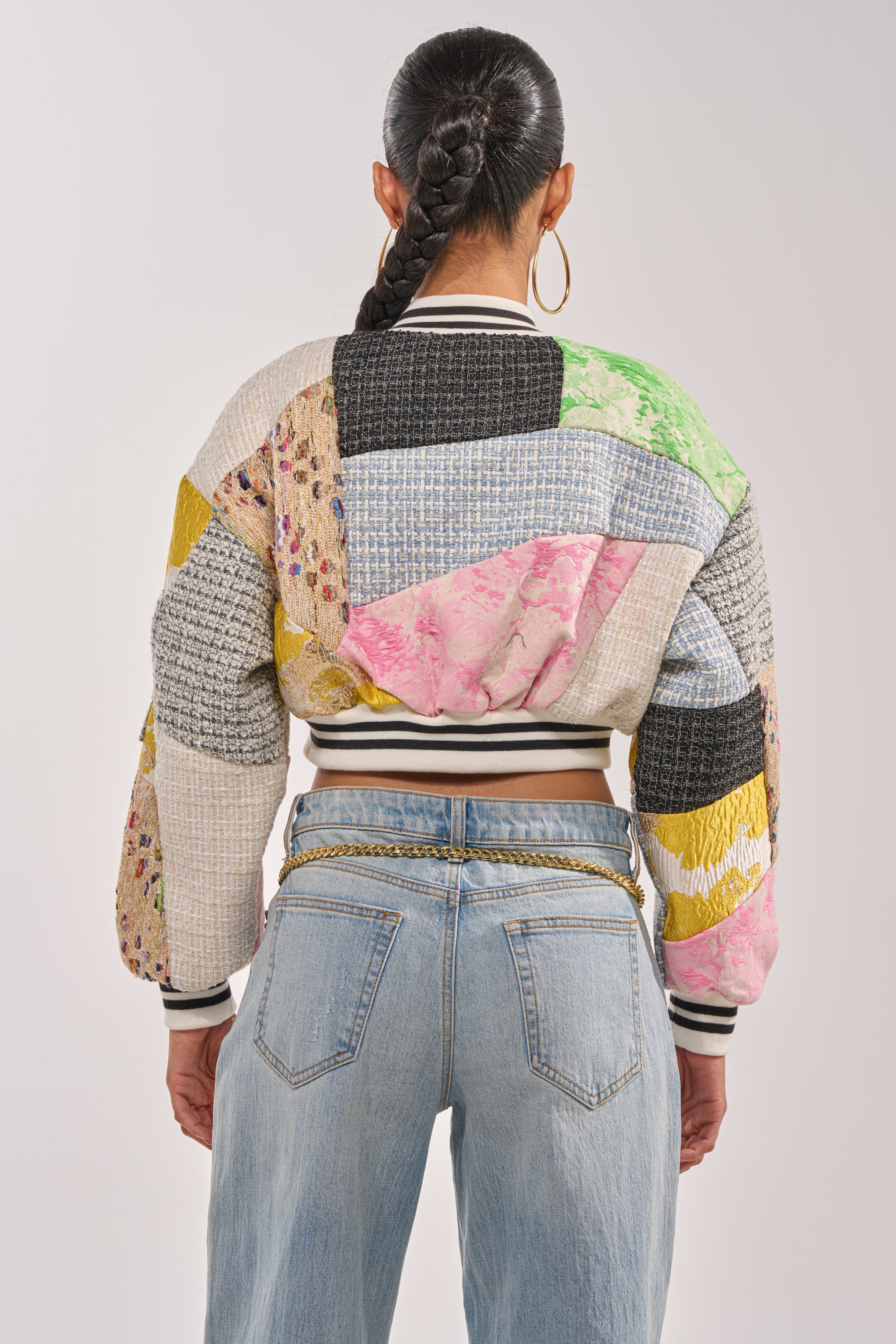 A woman with a long braid and hoop earrings wears the ESTELLE PATCHWORK BOMBER and light blue jeans with a chain belt, standing with her back to the camera against a plain background. 7