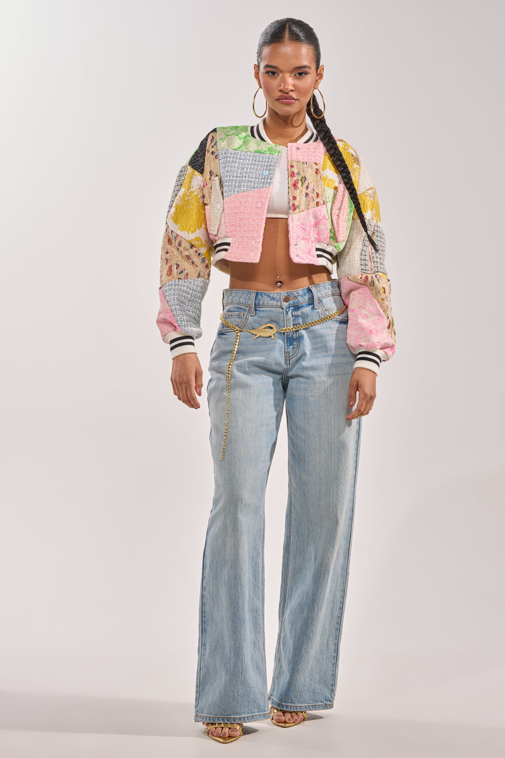 A woman stands facing forward, wearing the ESTELLE PATCHWORK BOMBER over a white crop top, paired with light blue wide-leg jeans with a gold chain, hoop earrings, and sandals against a plain light background. 8