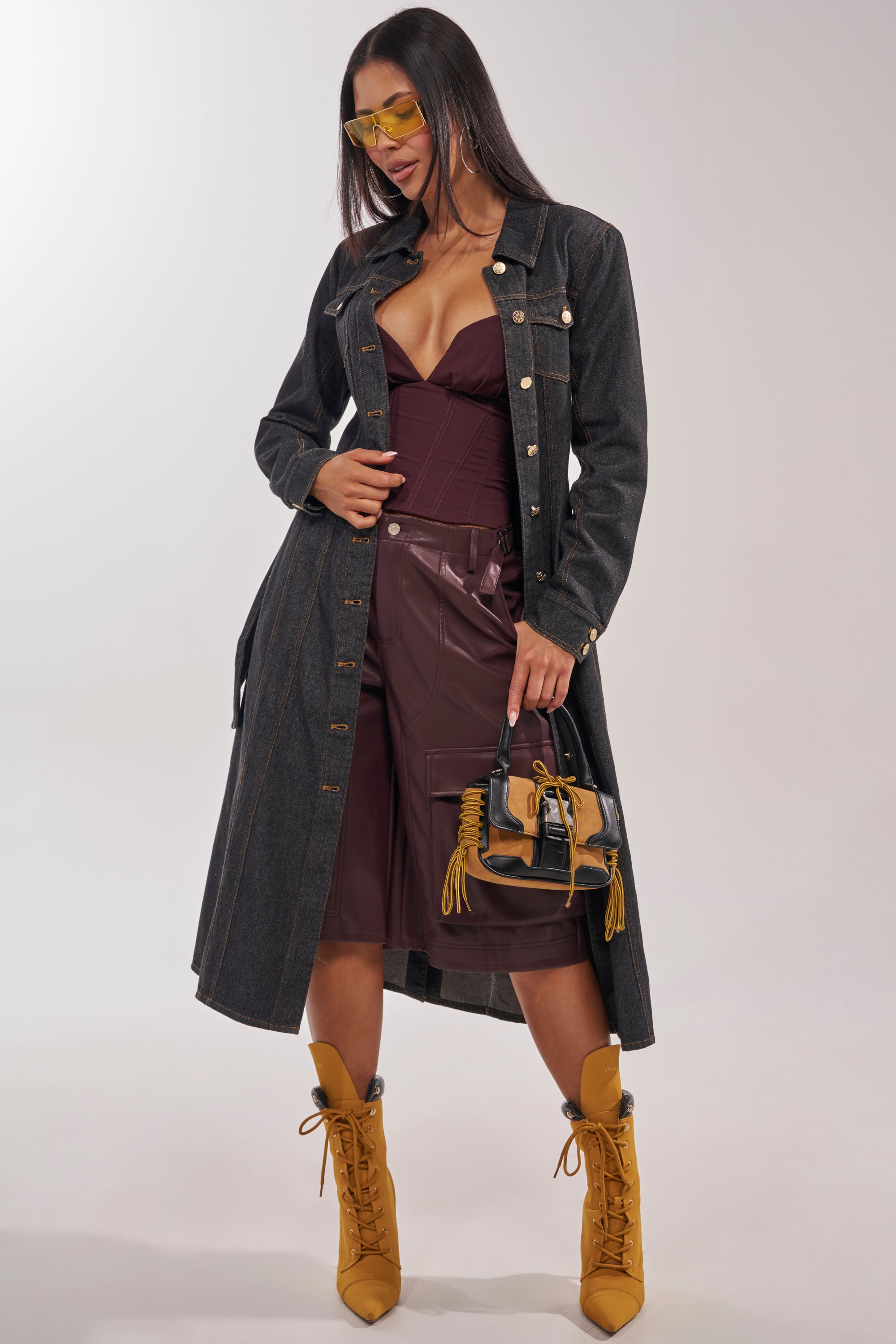 A stylish woman models the ON YOUR RADAR DENIM TRENCH over a burgundy bustier and matching leather shorts, paired with yellow heeled boots, yellow sunglasses, and a small handbag featuring yellow accents. 1