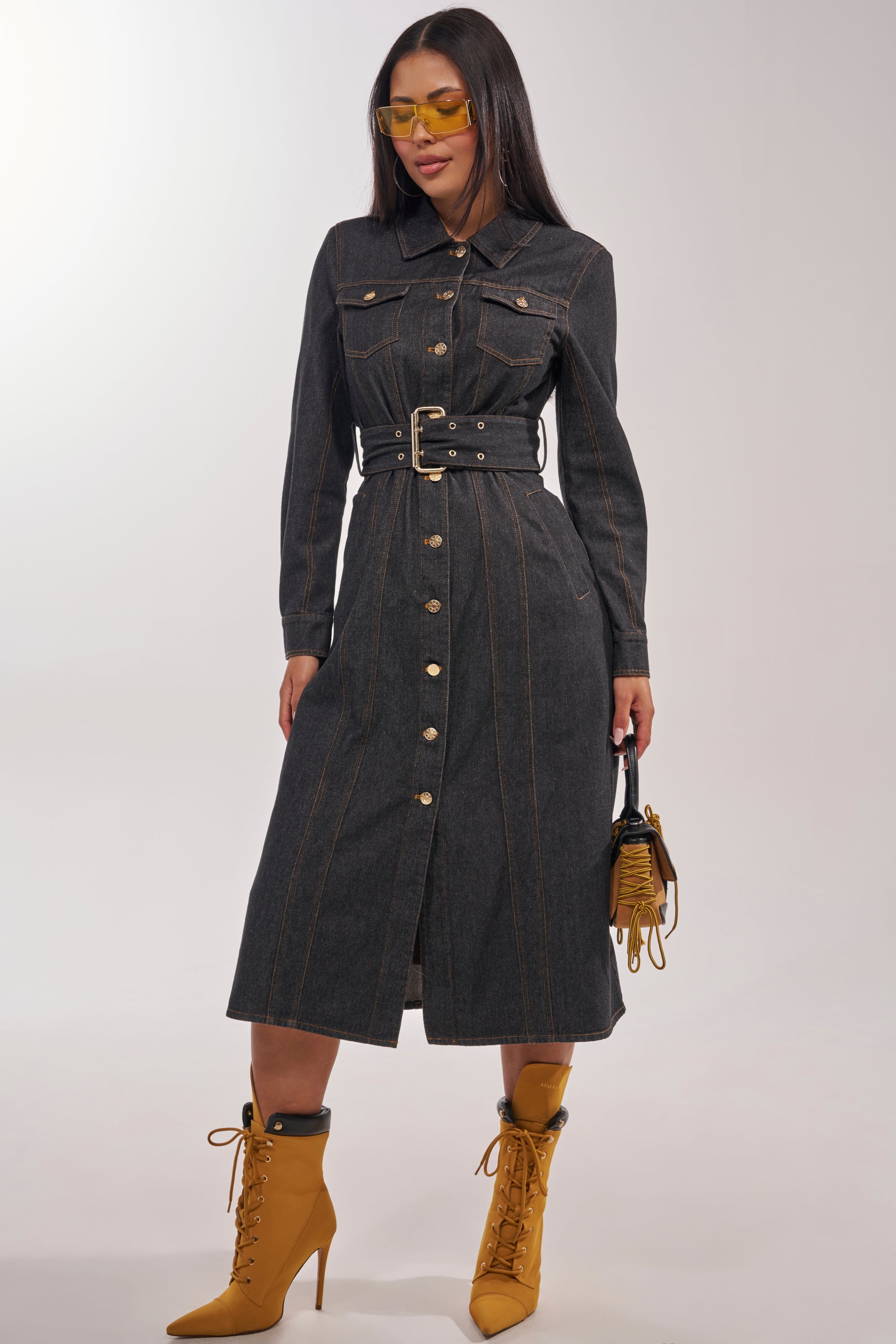 A woman wearing the ON YOUR RADAR DENIM TRENCH—a long-sleeve, belted, button-down dark-wash denim coat with gold stitching—paired with yellow heeled lace-up boots, yellow tinted sunglasses, and a small handbag. 0
