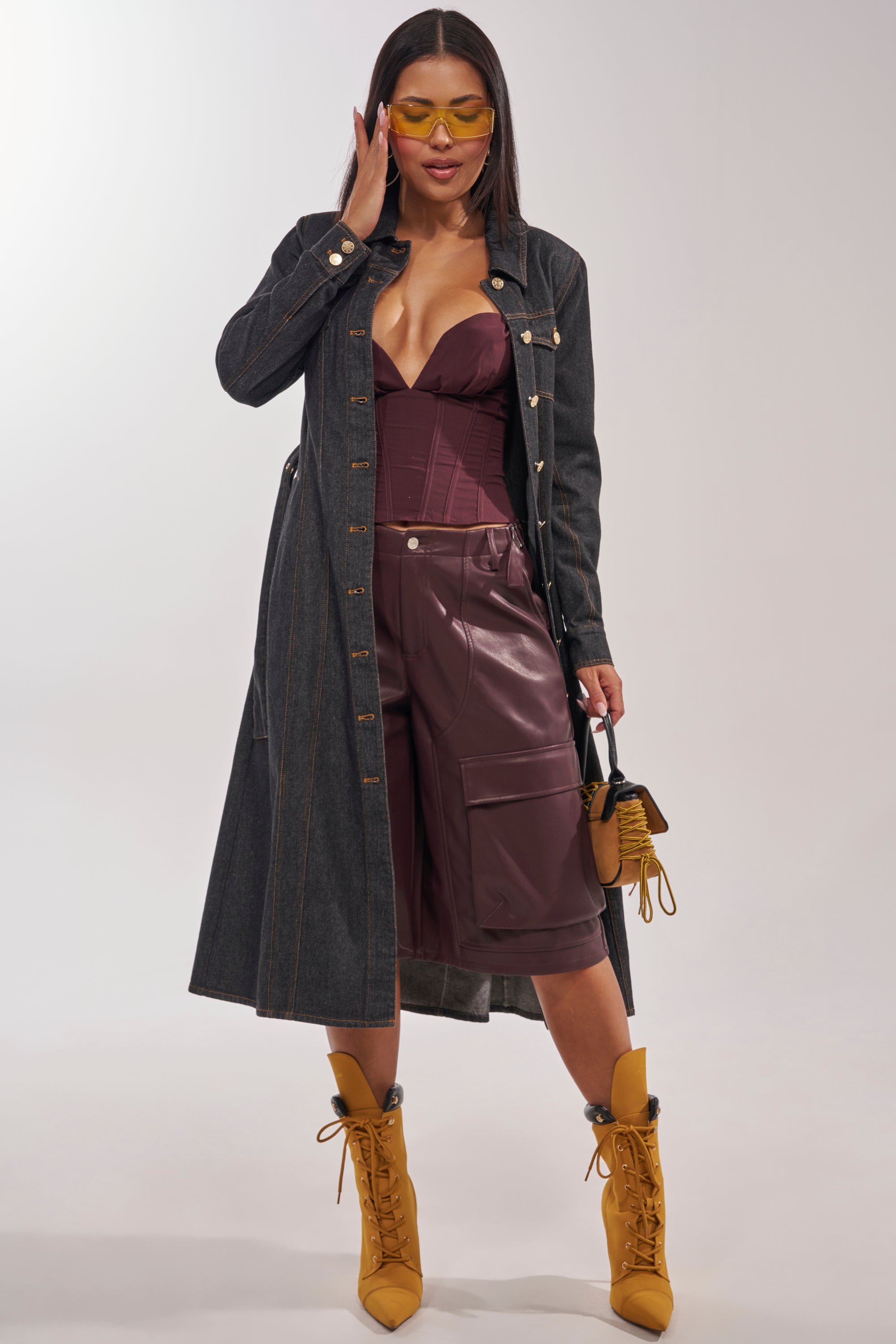 A woman poses confidently in the ON YOUR RADAR DENIM TRENCH over a burgundy bustier and matching leather shorts, styled with yellow boots, yellow-tinted sunglasses, and a small yellow handbag. 2