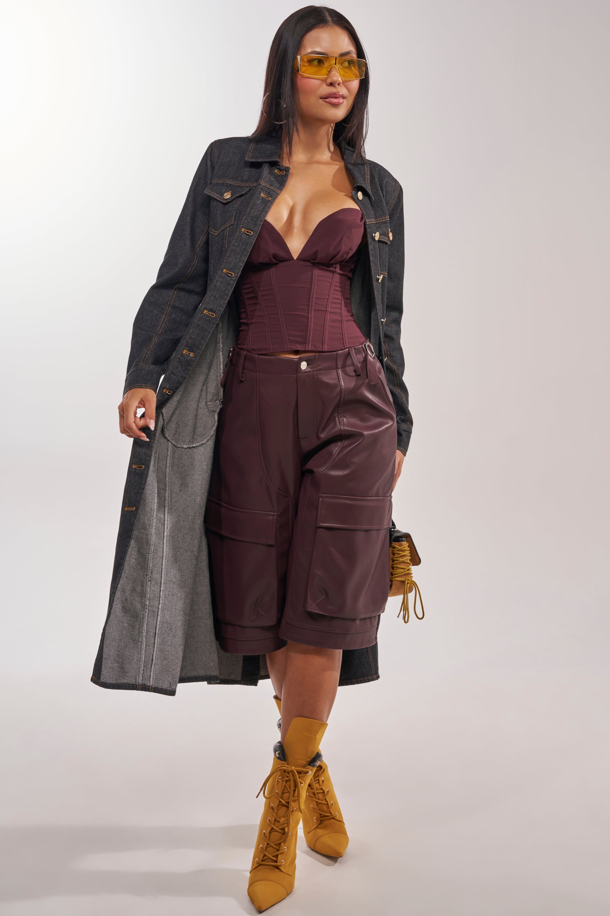 A woman in yellow sunglasses and the ON YOUR RADAR DENIM TRENCH pairs it with a burgundy bustier, matching knee-length cargo shorts, mustard lace-up boots, and a small yellow purse as she walks confidently. 3