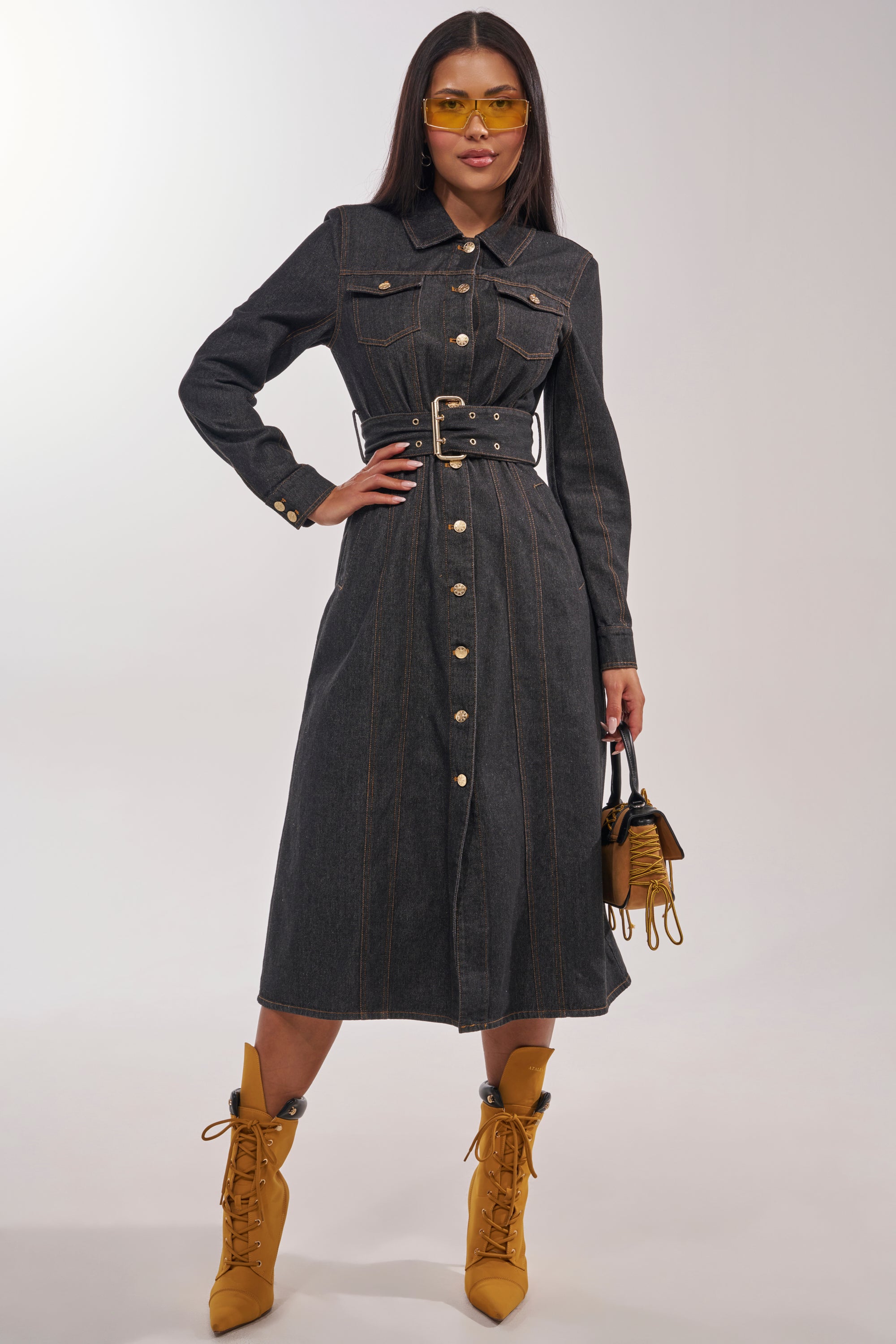 A woman confidently models the ON YOUR RADAR DENIM TRENCH as a dress, pairing the belted long-sleeve coat with yellow boots, yellow-tinted sunglasses, and a small brown handbag for a chic, coordinated look. 4