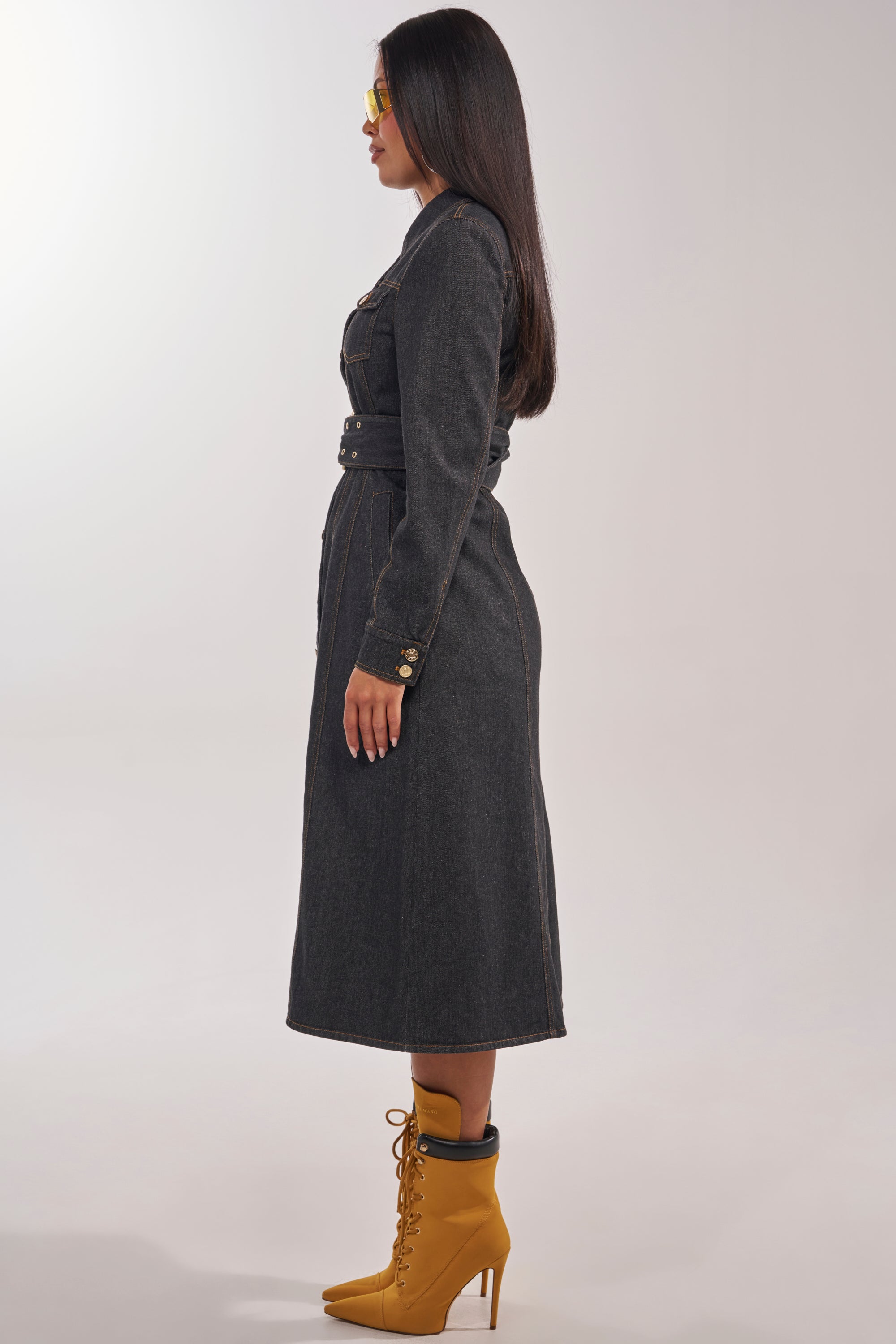 A woman stands in profile against a plain light background wearing the ON YOUR RADAR DENIM TRENCH with gold buttons, yellow-tinted sunglasses, and mustard yellow lace-up ankle boots. 5