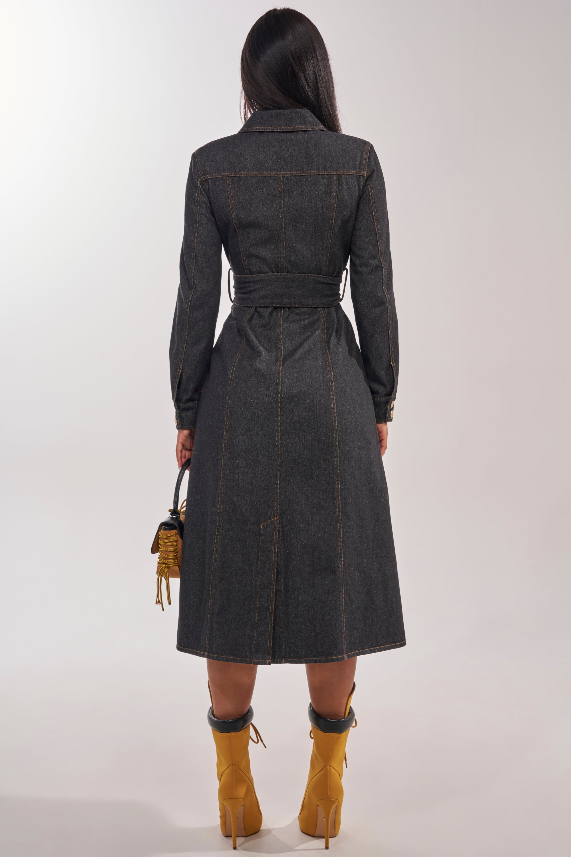 A woman faces away, wearing the ON YOUR RADAR DENIM TRENCH with yellow high-heeled boots and a small yellow handbag. Her long hair cascades down against a simple, light backdrop. 6