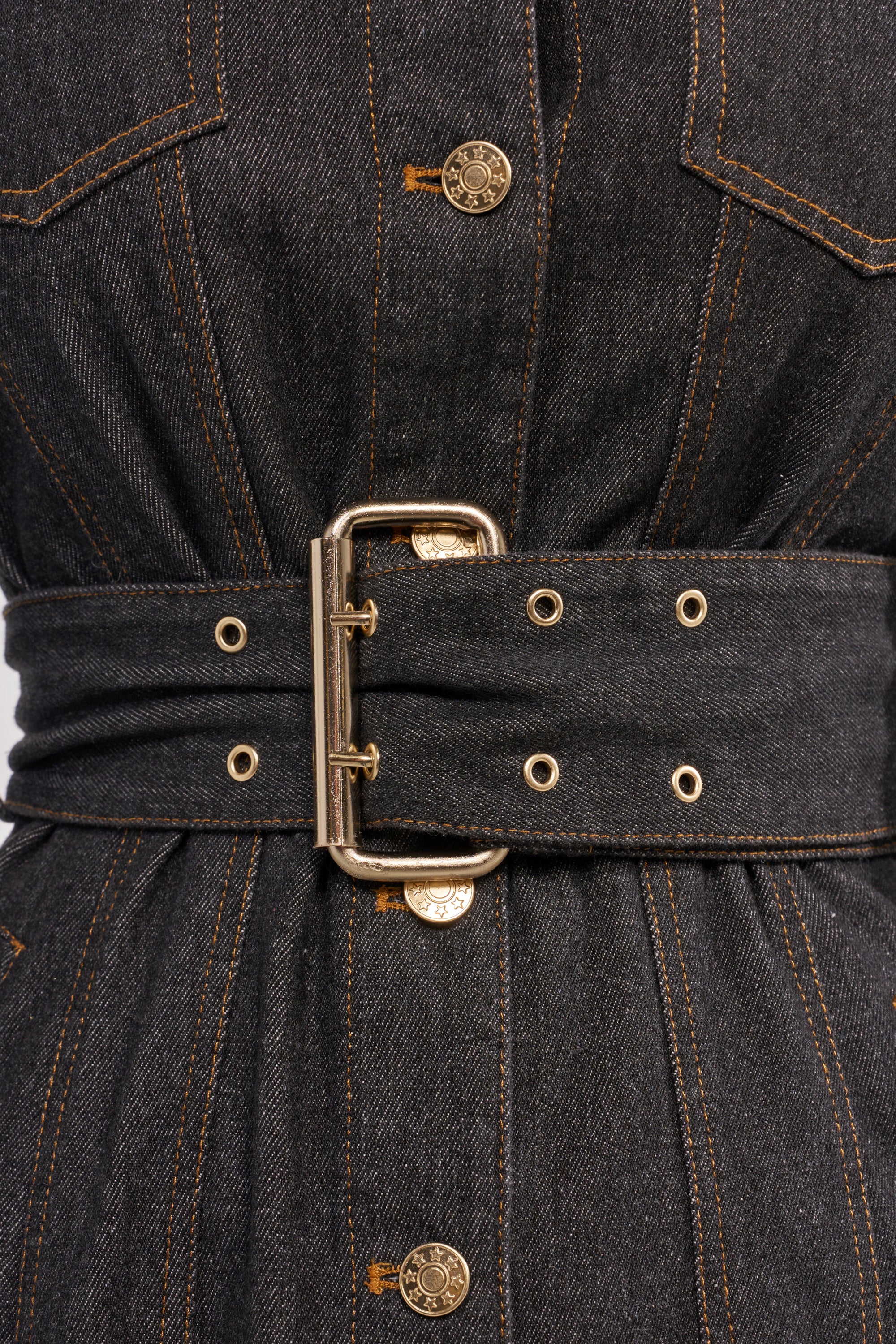 Close-up of the ON YOUR RADAR DENIM TRENCH: a dark-wash cotton denim dress with gold buttons, gold stitching, a wide matching belt with a large gold buckle and eyelets, plus front flap pockets. 7