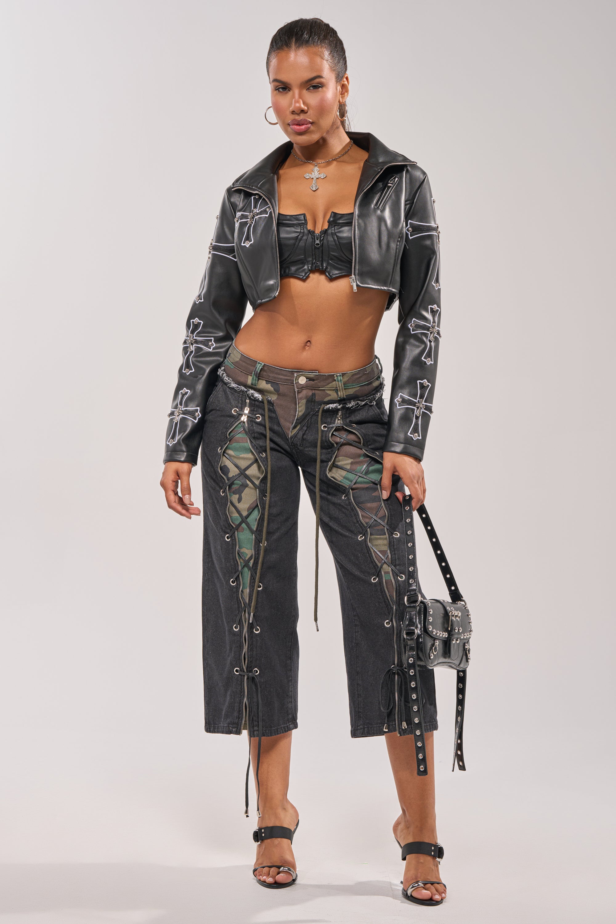 A woman stands confidently in the SIN CITY CROPPED MOTO with white stitching, a black bandeau, lace-up black camo pants, heeled sandals, a cross necklace, and a studded black bag. 11