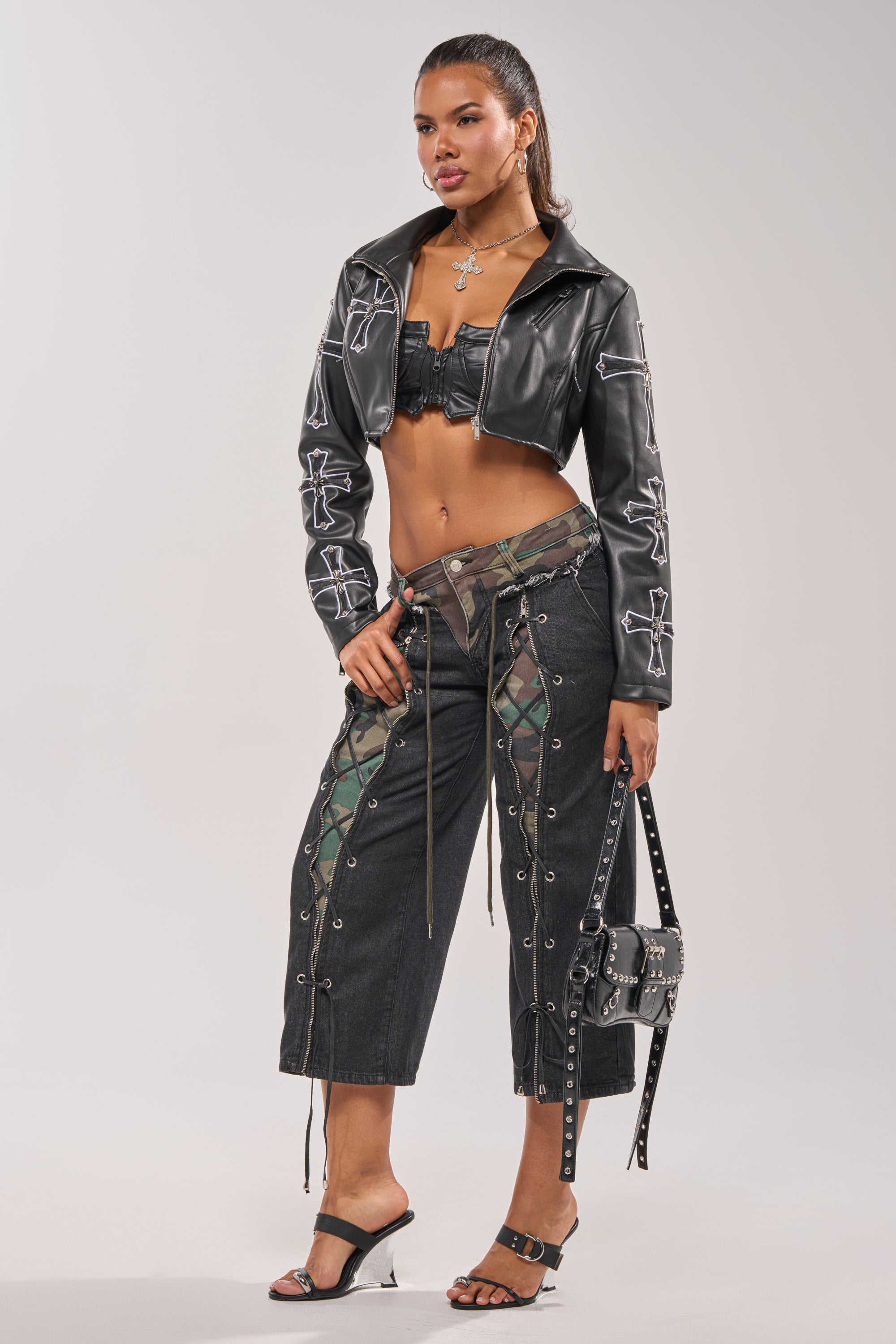 A woman poses confidently in the SIN CITY CROPPED MOTO leather jacket with white stitching, pairing it with a black crop top, dark wide-leg lace-up camo pants, heeled sandals, cross necklaces, and a studded handbag. 12