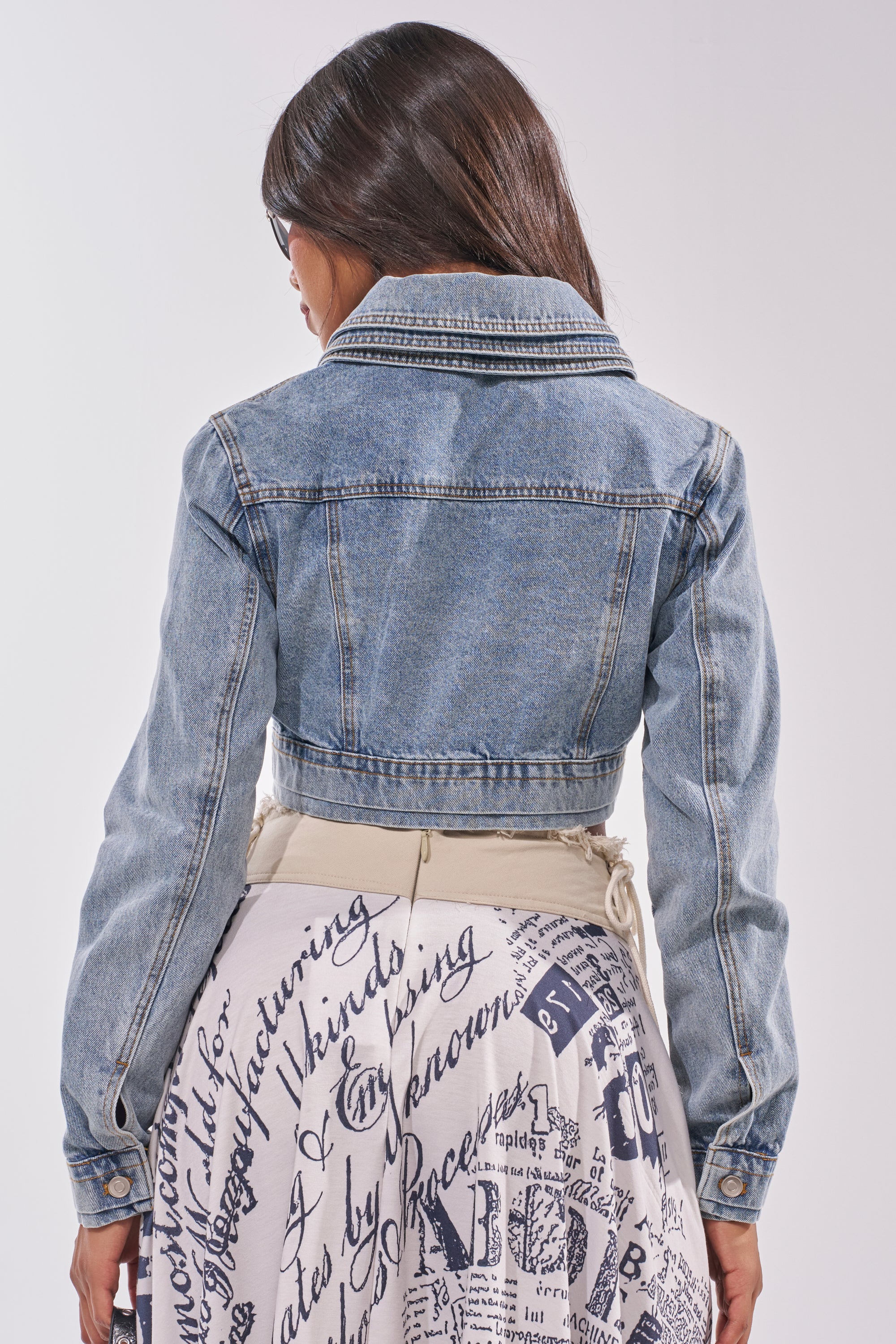 A woman with long brown hair, seen from behind, wears the NO REPEATS LAYERED DENIM JACKET with a layered collar and a white skirt with bold black text and graphics. The background is plain and light-colored. 7