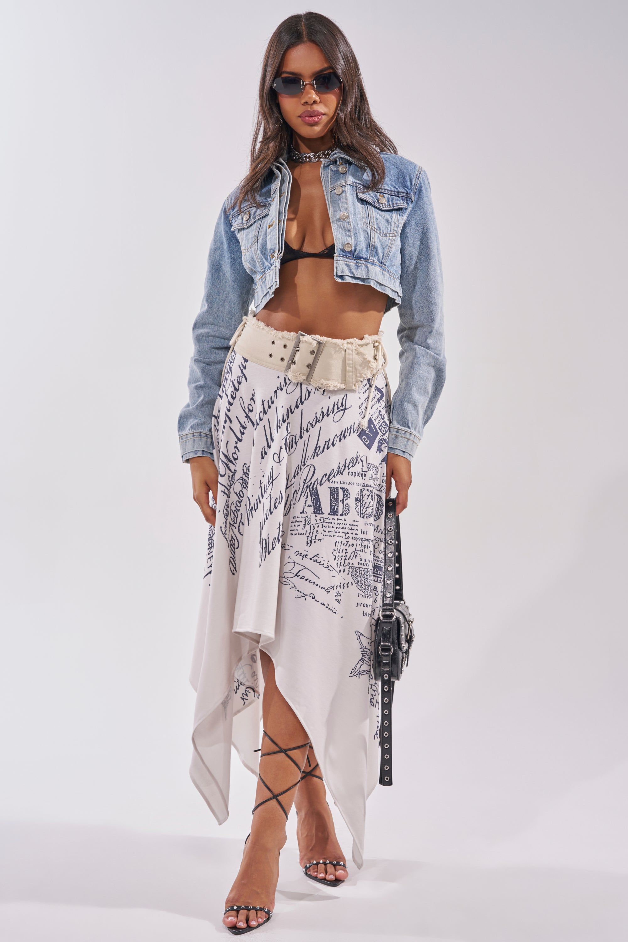A fashion-forward woman styles the NO REPEATS LAYERED DENIM JACKET over a black bralette, pairing it with an asymmetrical white skirt featuring navy script, strappy heels, sunglasses, and a black handbag. 8