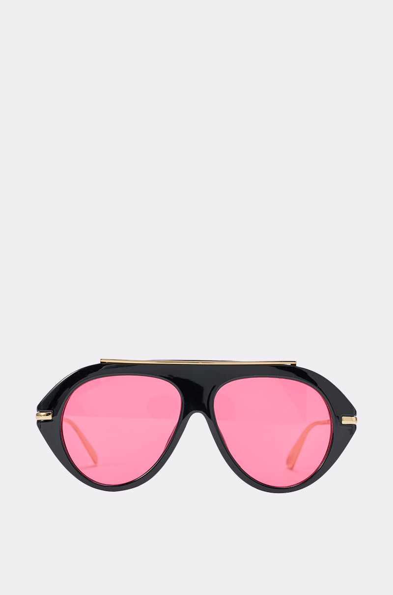 The CRIMSON VICE AVIATOR SUNGLASSES feature a bold black frame with pink-tinted lenses and striking gold accents, showcased on a simple light gray background. This modern, angular design is inspired by oversized aviator styles.
