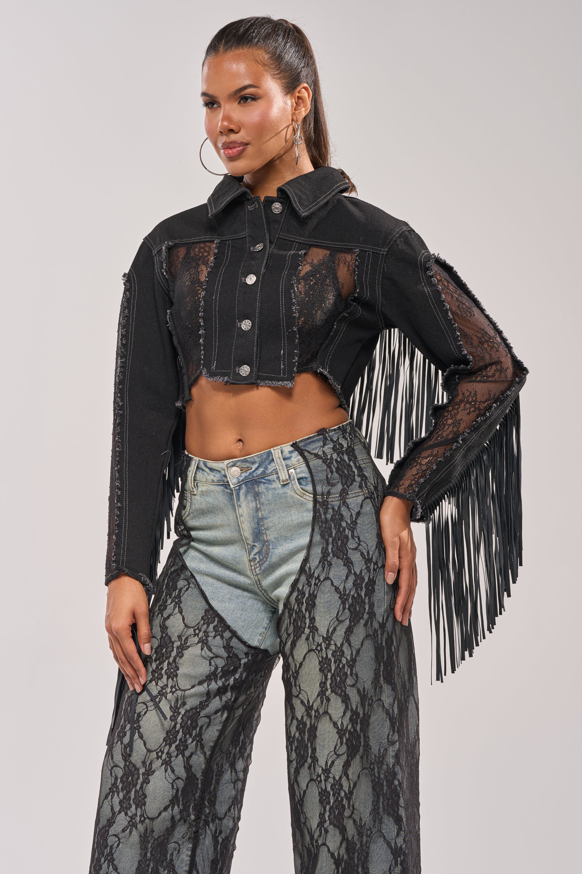 A woman stands confidently against a plain background in the LACED AND LOADED DENIM JACKET—a cropped black festival piece with lace panels and fringe sleeves—styled with unique high-waisted pants featuring one lace and one denim leg. 0