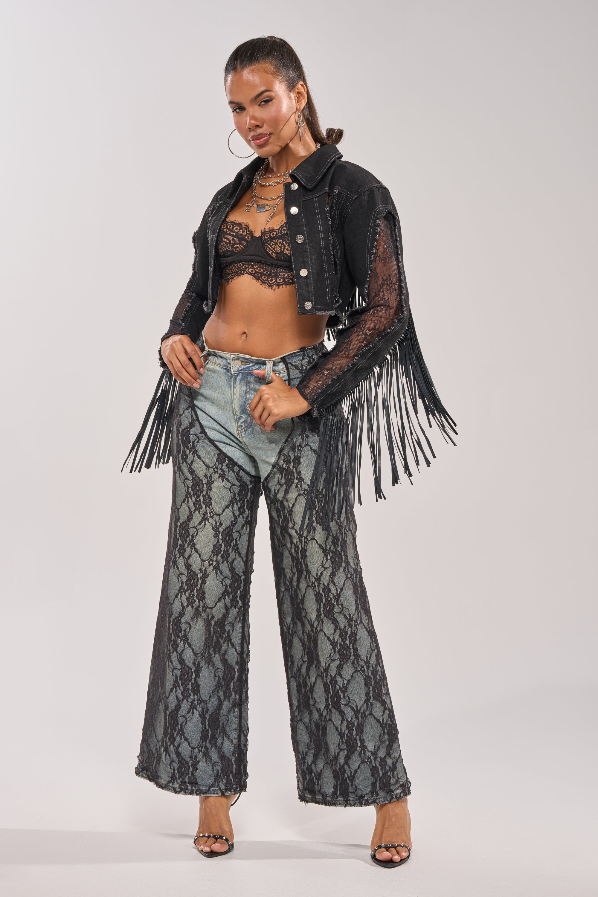 A woman poses in the LACED AND LOADED DENIM JACKET, a black lace bralette, wide-leg jeans with black lace overlay, hoop earrings, layered necklaces, and black heeled sandals. 9