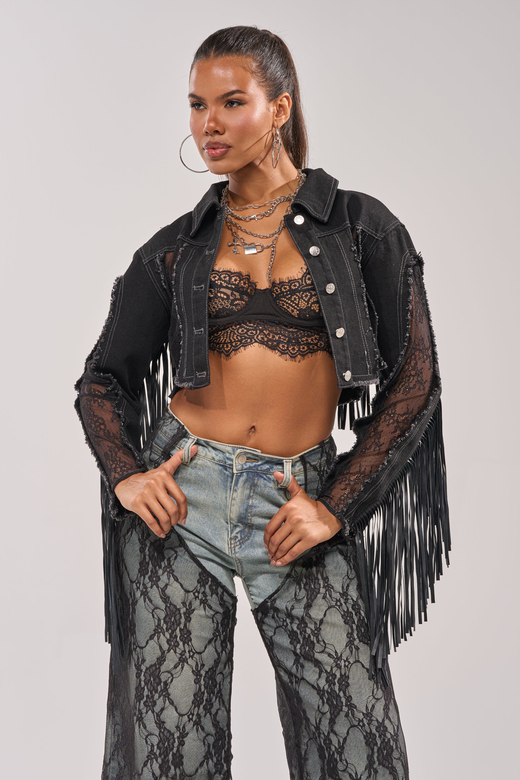 A woman poses with her hands on her hips wearing the LACED AND LOADED DENIM JACKET layered over a black lace bralette, hoop earrings, high-waisted patterned jeans, and layered necklaces against a plain background. 1