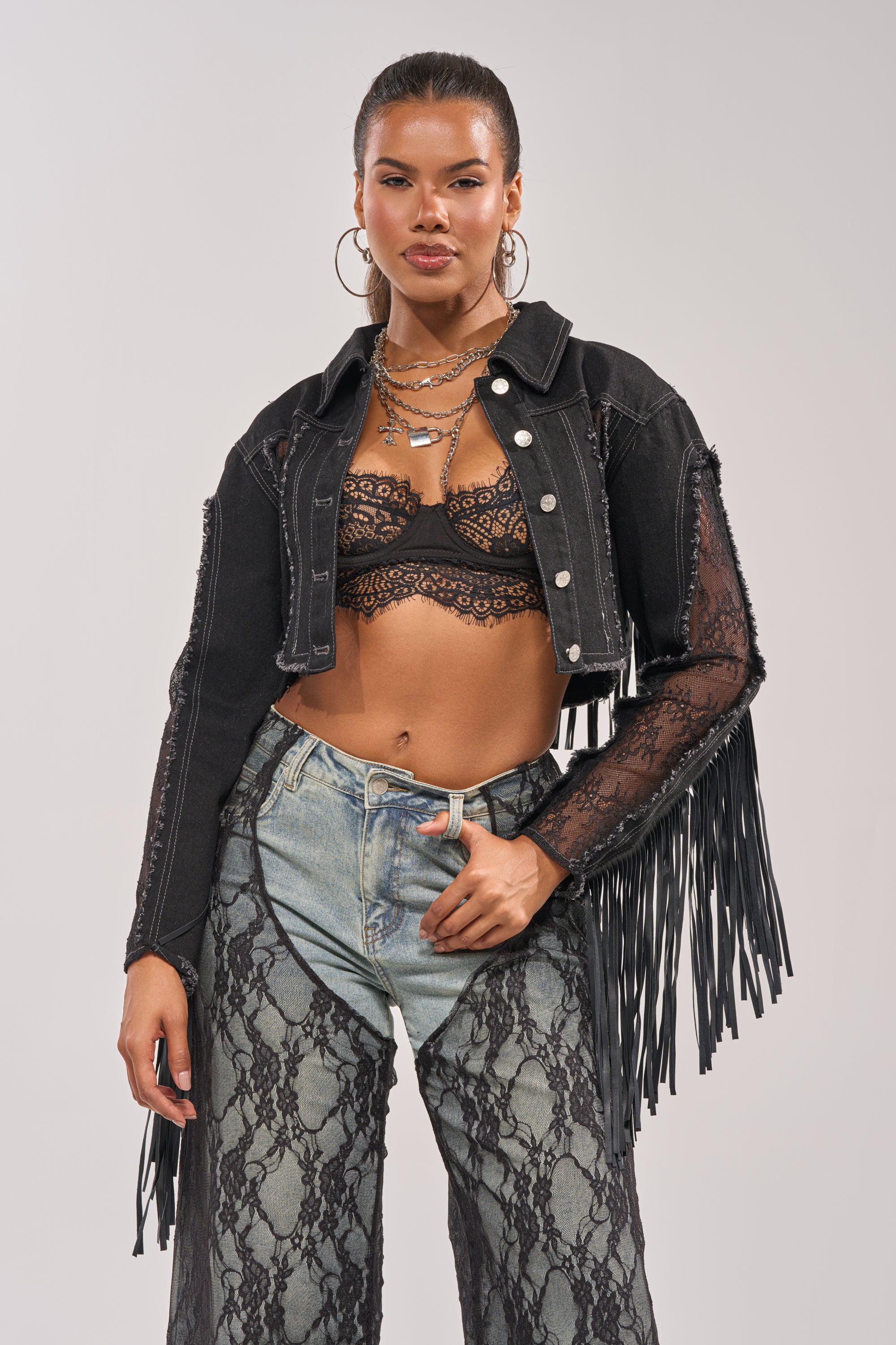 A woman confidently wears the LACED AND LOADED DENIM JACKET—black denim with fringe and lace sleeves—over a black lace bralette and high-waisted, light-wash jeans with lace overlay, styled with layered necklaces and hoop earrings for a western festival look. 2