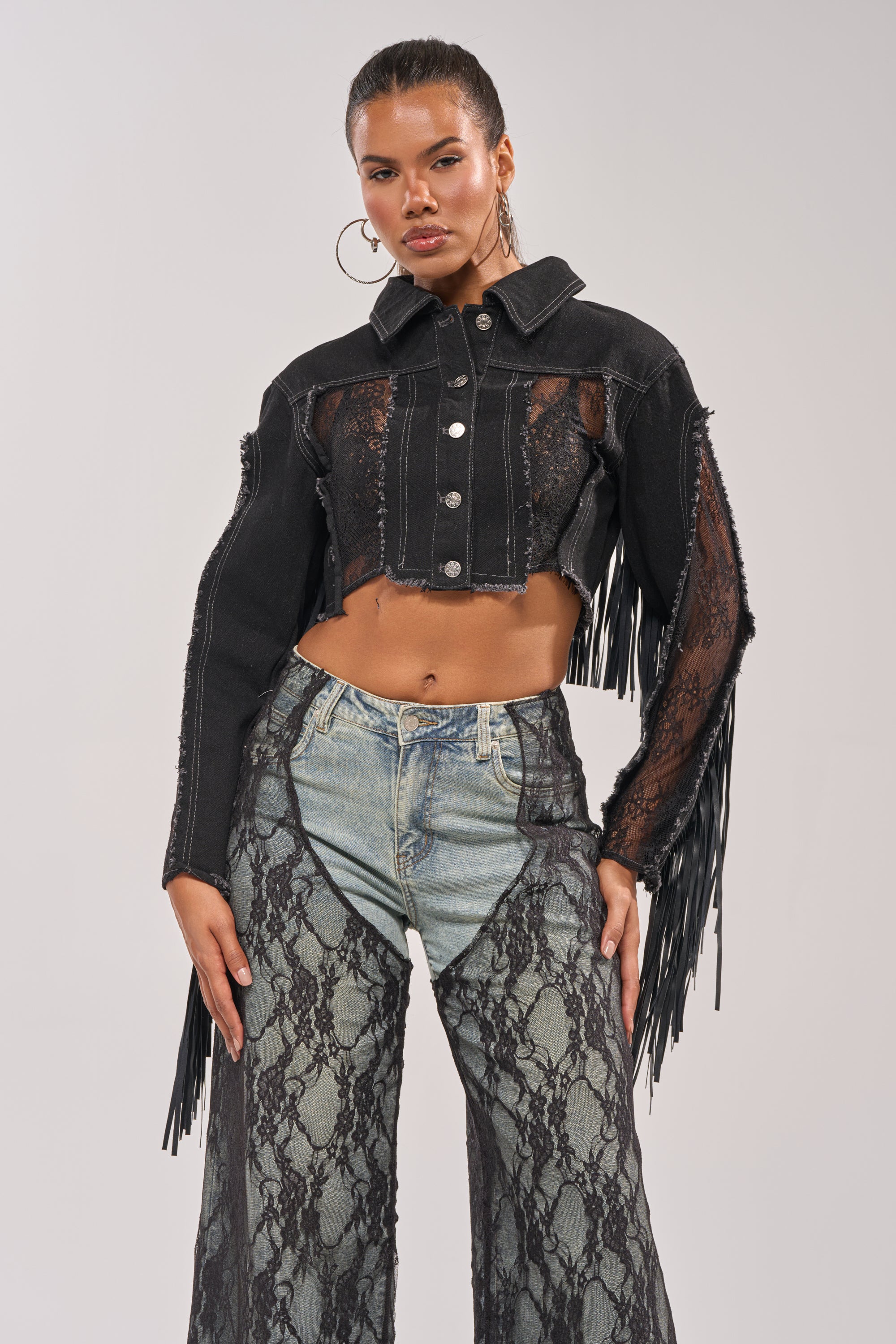 A woman models the LACED AND LOADED DENIM JACKET, a black cropped western festival piece with fringe and lace details, styled with high-waisted pants and hoop earrings for a bold, confident look. 3
