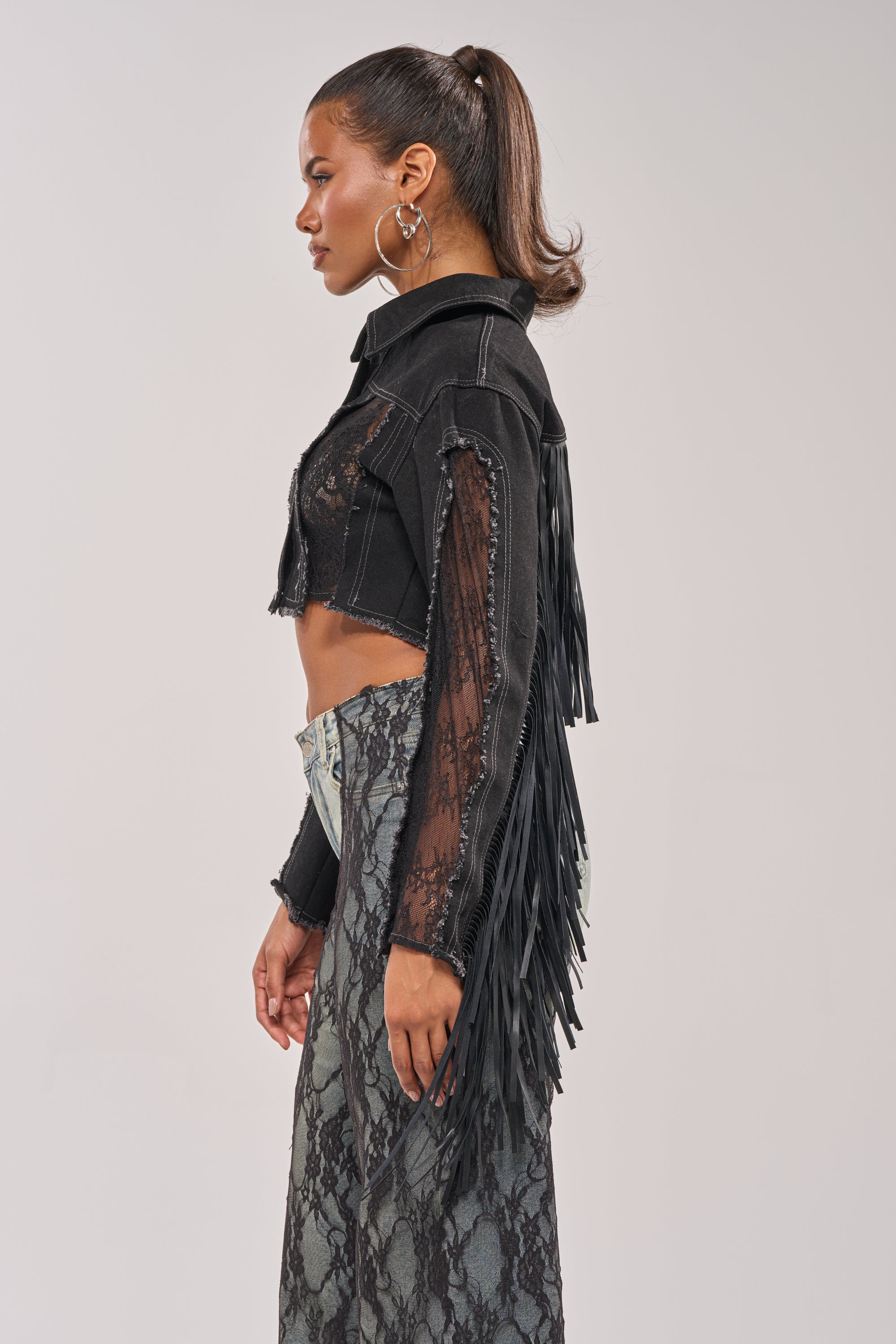 A woman facing sideways wears the LACED AND LOADED DENIM JACKET with lace sleeves, hoop earrings, and high-waisted lace pants. Her hair is styled in a high ponytail against a plain light gray background. 4