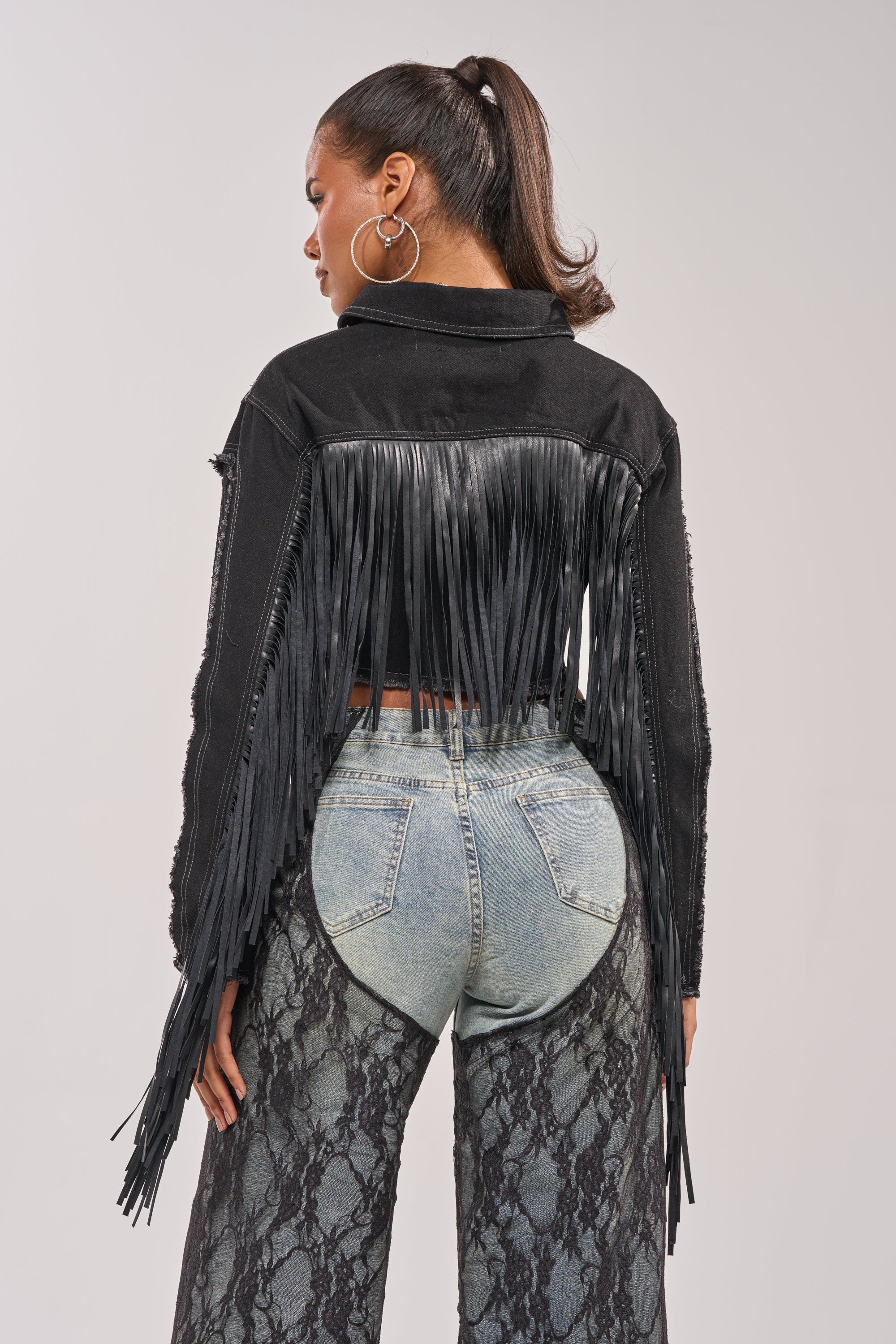 A woman with a ponytail, hoop earrings, and high-waisted snakeskin jeans is seen from behind wearing the LACED AND LOADED DENIM JACKET, a black western style with long fringe on the back and sleeves. 6