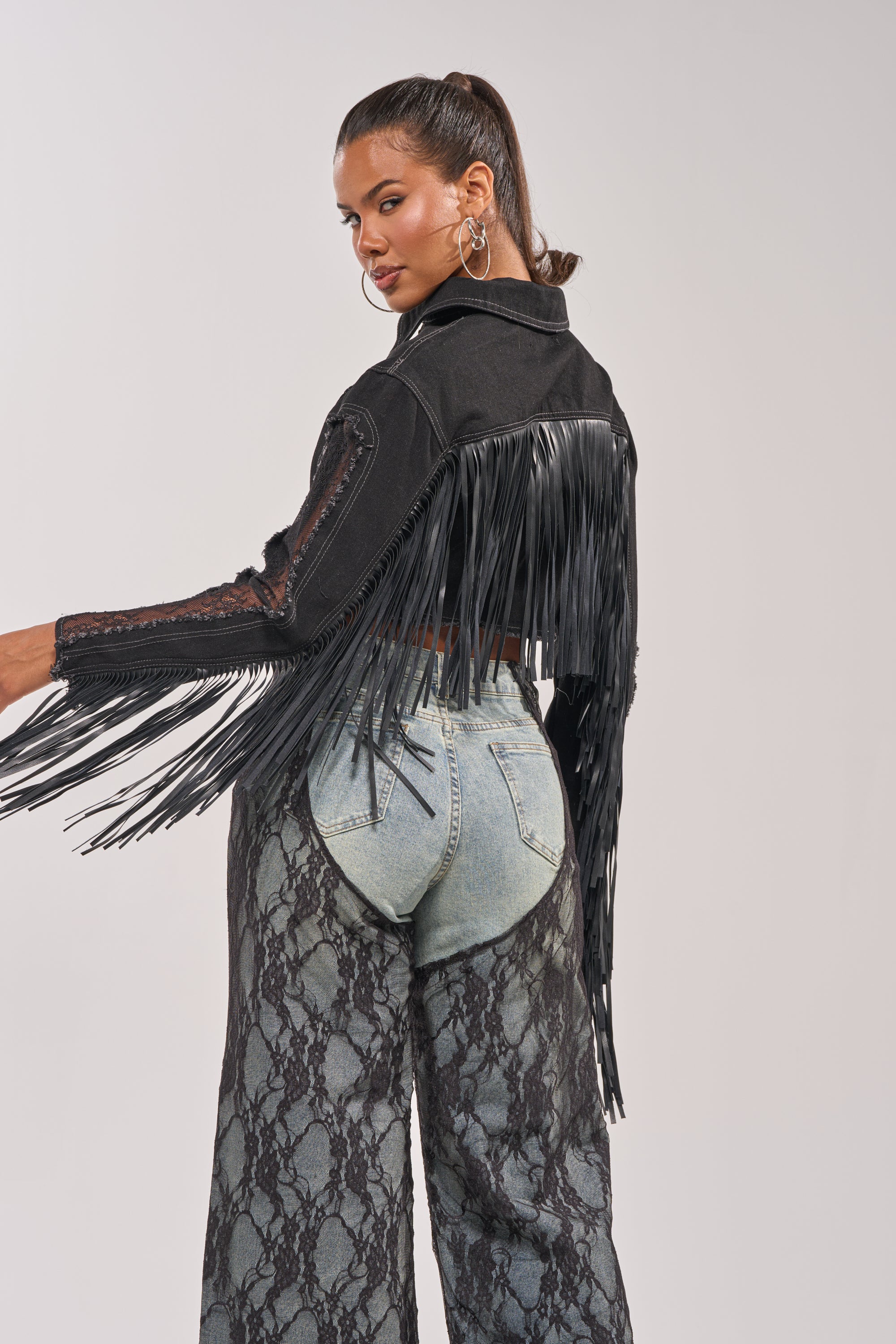 A woman poses in the LACED AND LOADED DENIM JACKET with long fringe and high-waisted, light denim snakeskin print jeans. Her hair is styled in a high ponytail, and she accessorizes with large hoop earrings. 7