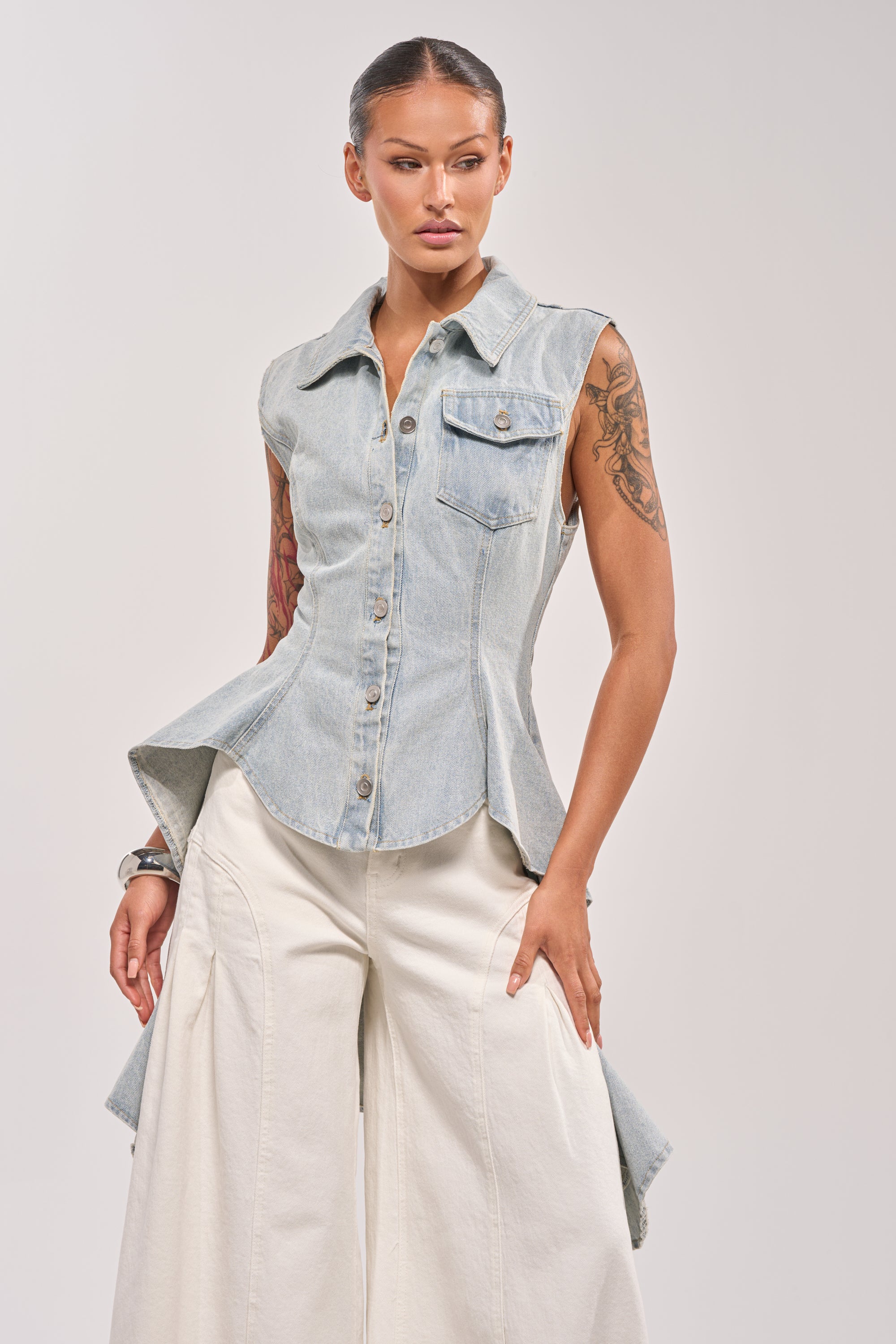 A woman with slicked-back hair and arm tattoos poses against a light background, wearing the HIGH LOW DENIM VEST in a light wash with an asymmetrical hem, paired with high-waisted wide-leg white pants. 0