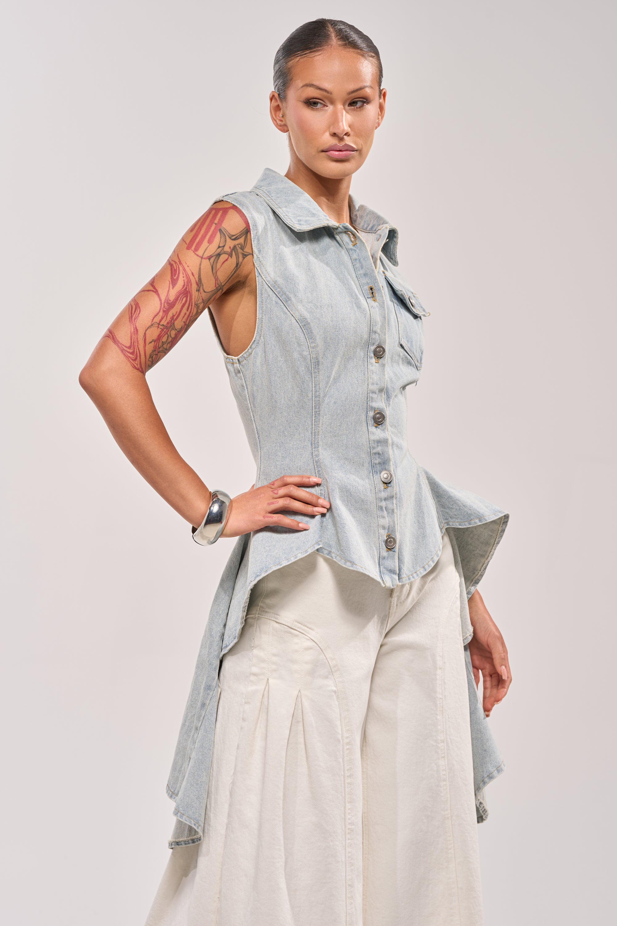 A woman with a red floral tattoo on her arm models the HIGH LOW DENIM VEST, featuring a light wash and high-low hem, styled over wide cream pleated pants as she poses confidently against a plain background. 1