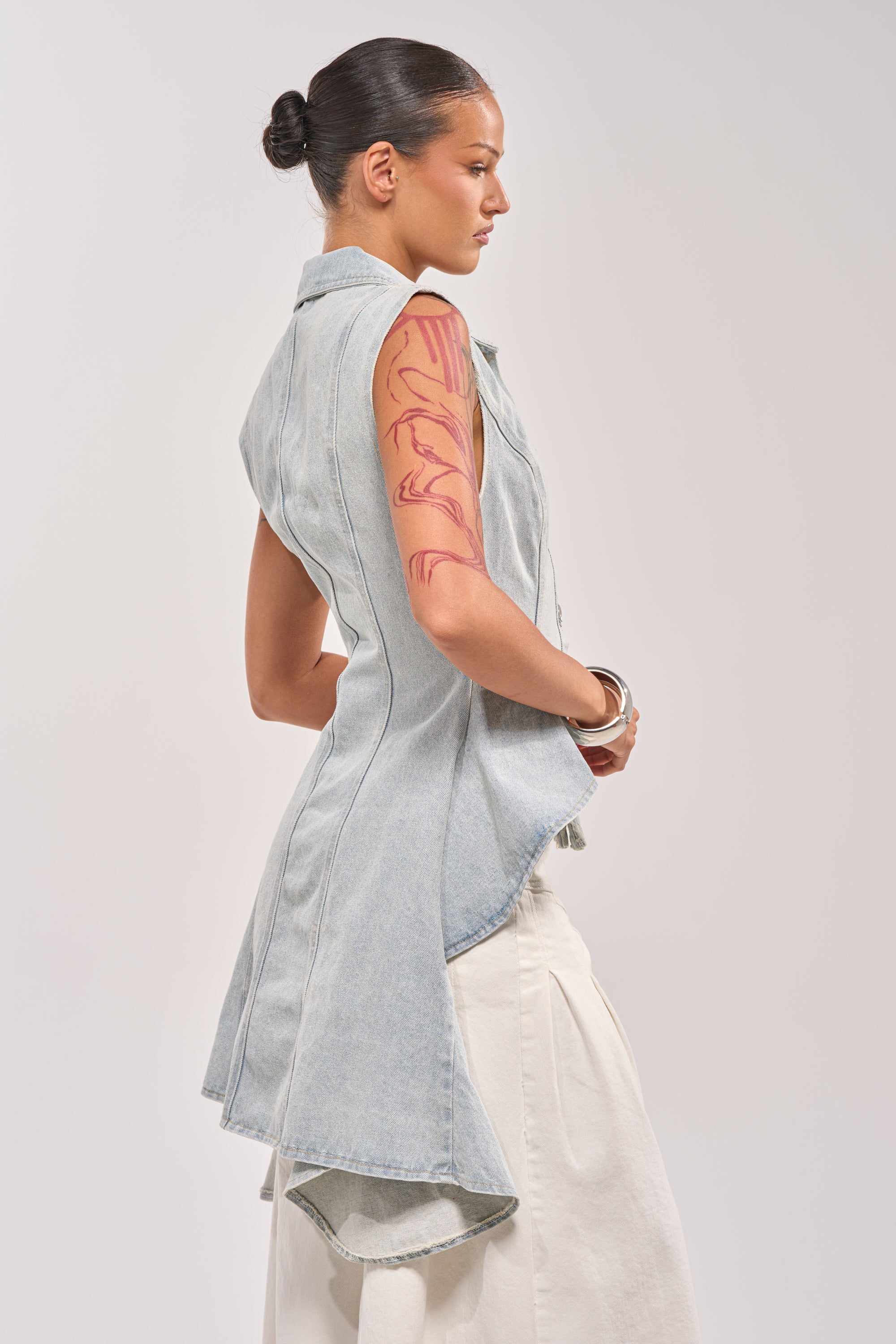 A woman with dark hair in a bun models the HIGH LOW DENIM VEST, a sleeveless light-wash design with a high-low hem, worn over a white skirt. Shown in profile, she displays a red abstract tattoo on her upper arm against a plain backdrop. 2