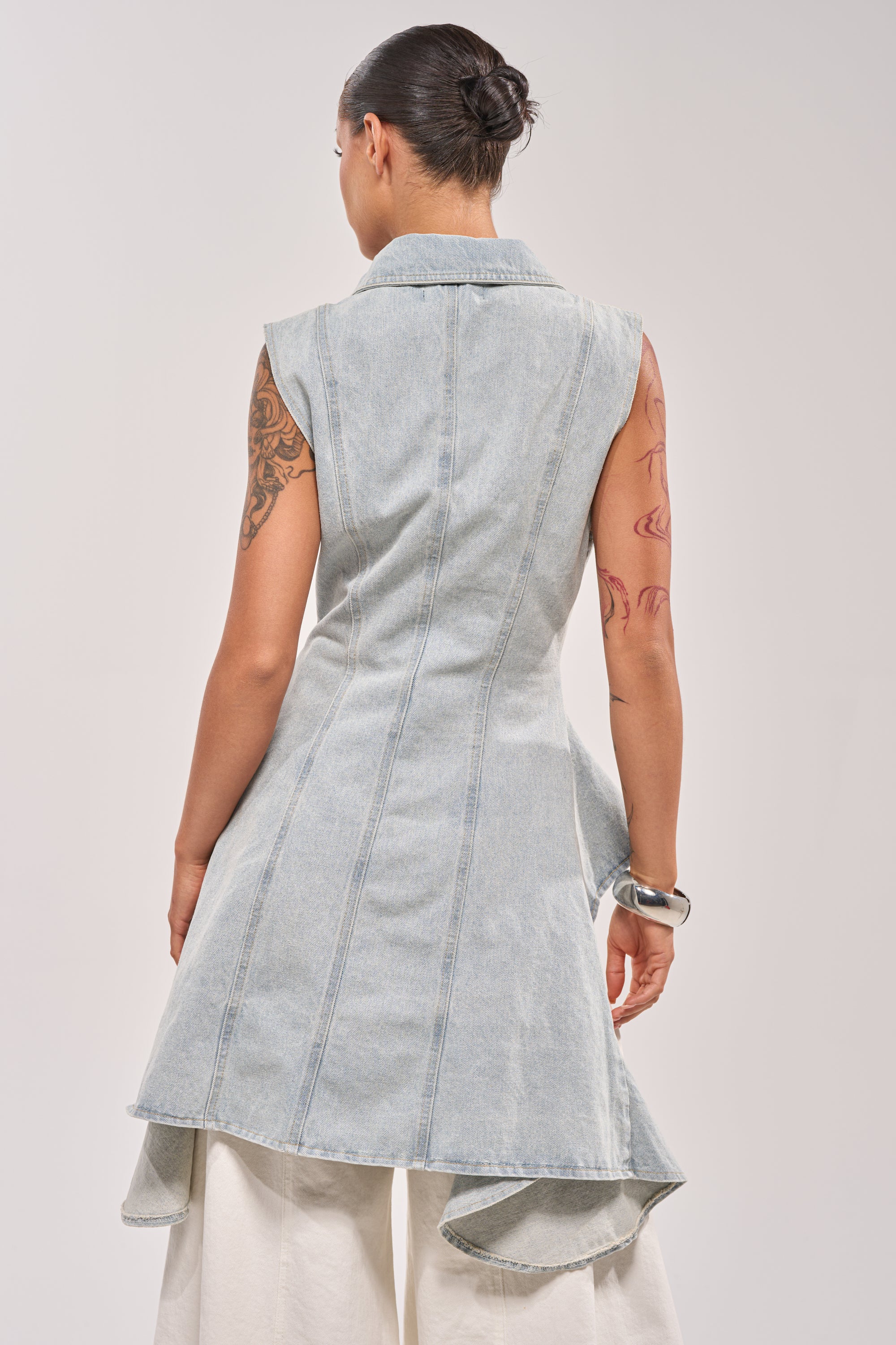 A woman with her hair in a bun and tattoos on her left arm stands with her back to the camera, wearing the HIGH LOW DENIM VEST— a sleeveless, light wash denim piece with a flared hem layered over white wide-leg pants. 3