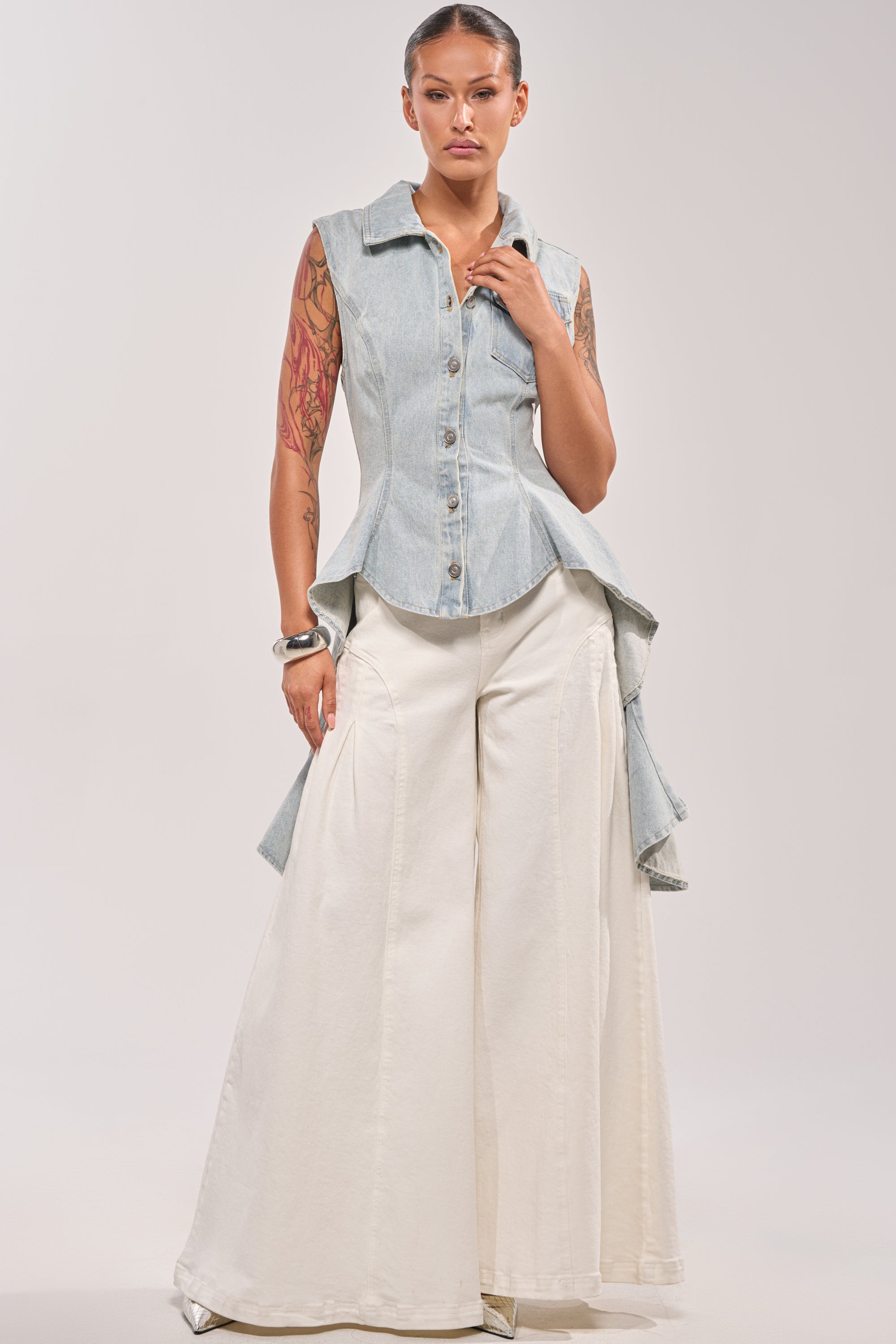 A tattooed woman wears the HIGH LOW DENIM VEST and wide-leg, high-waisted white pants. She stands facing forward, one hand at her side and the other touching her collar. 4