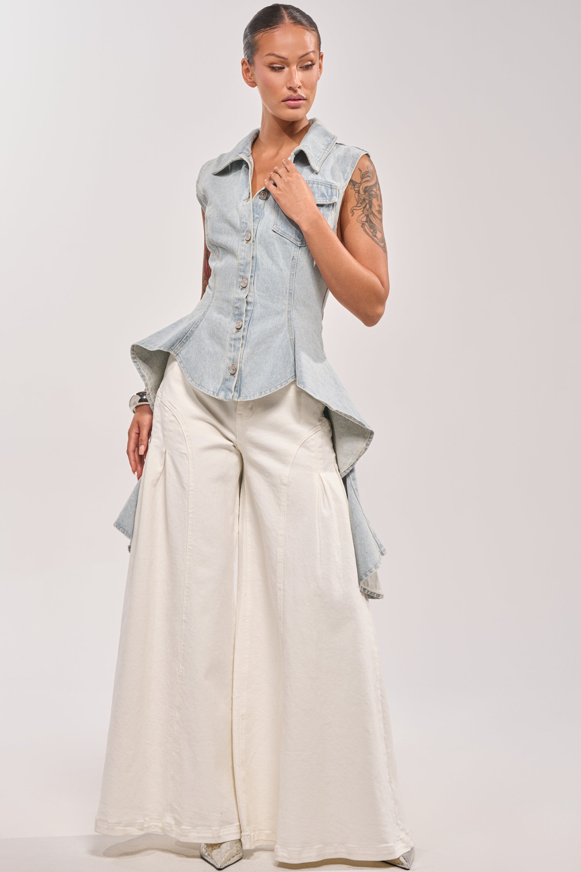 A woman poses in the HIGH LOW DENIM VEST, a sleeveless light wash design with a collar and flared tails, styled with high-waisted wide-leg white pants and pointed shoes against a plain light background. 5
