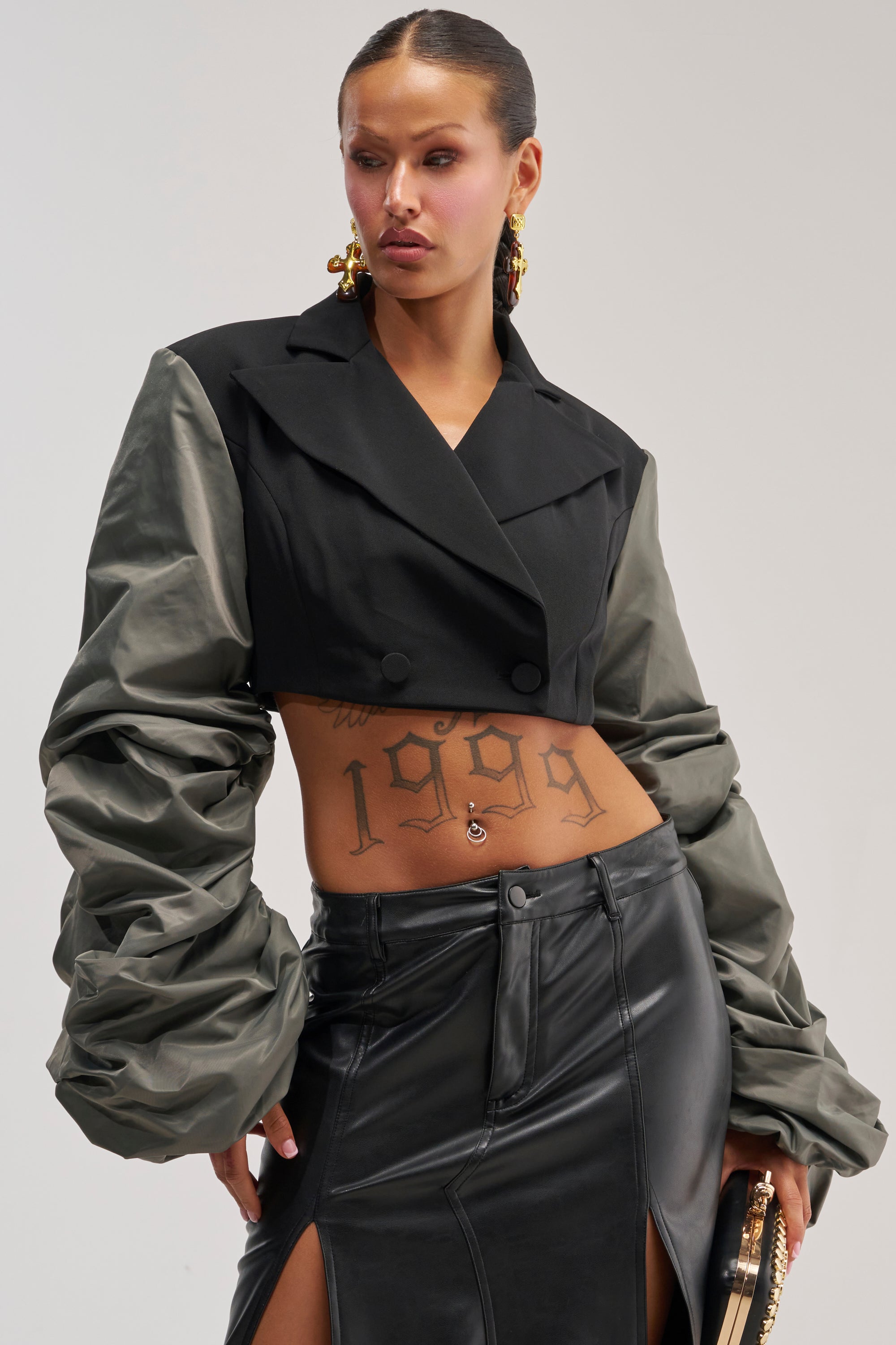 A person exudes confidence in the MIXED FEELINGS RUCHED SLEEVE BLAZER, its black cropped design and distinctive gray ruched sleeves creating a striking layered effect. Styled with a black leather skirt featuring thigh-high slits and gold accessories, the outfit draws attention to visible tattoos—including "1999" on their lower torso—for an unapologetically bold statement look.
