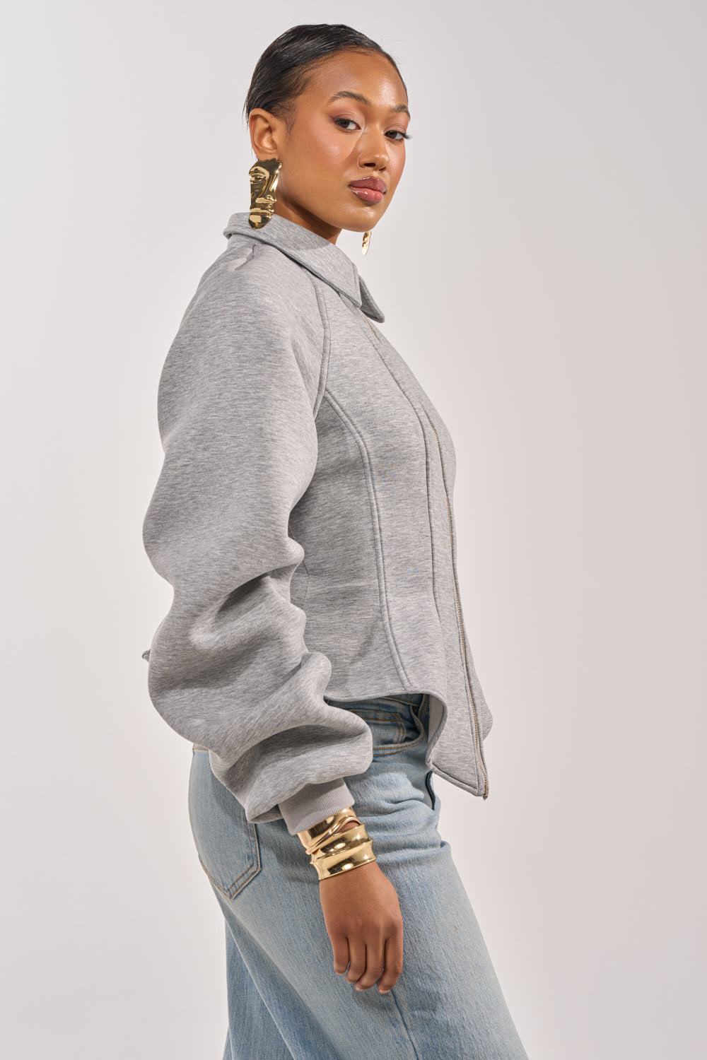 A woman poses confidently in the MONEY TALKS NEOPRENE JACKET, a light gray structured zip-up with puffy sleeves, paired with light blue jeans. She accessorizes with large gold earrings and stacked gold bracelets. 3