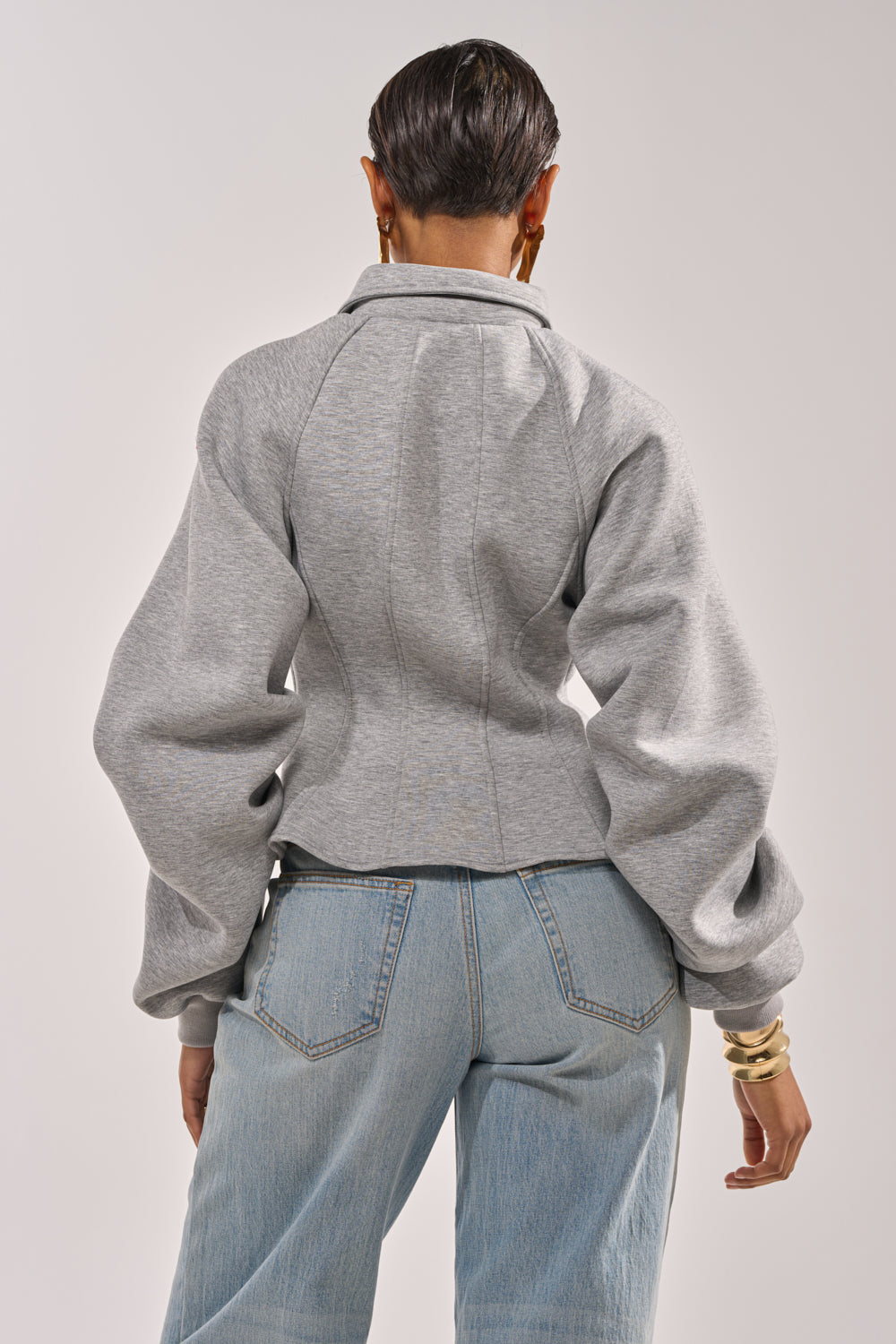 A person with short hair is seen from behind, wearing a fitted gray MONEY TALKS NEOPRENE JACKET with puffed sleeves and light blue jeans, standing against a plain background. 4