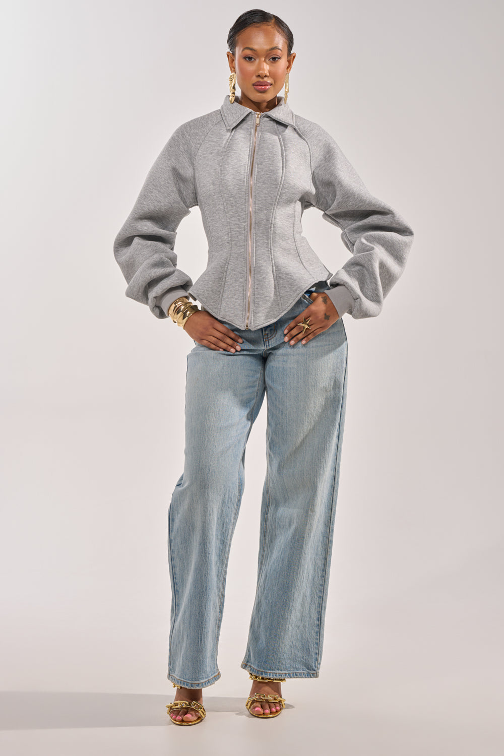 A woman stands confidently in the gray MONEY TALKS NEOPRENE JACKET with puffed sleeves, styled with light blue wide-leg jeans, gold hoop earrings, layered bracelets, and open-toe heels against a plain background. 5