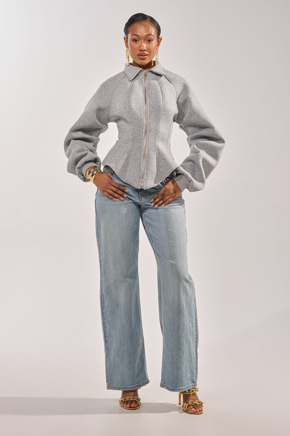 A woman faces forward, confidently wearing the MONEY TALKS NEOPRENE JACKET with puffed sleeves, light blue flared jeans, gold heels, and jewelry against a plain white background. 6