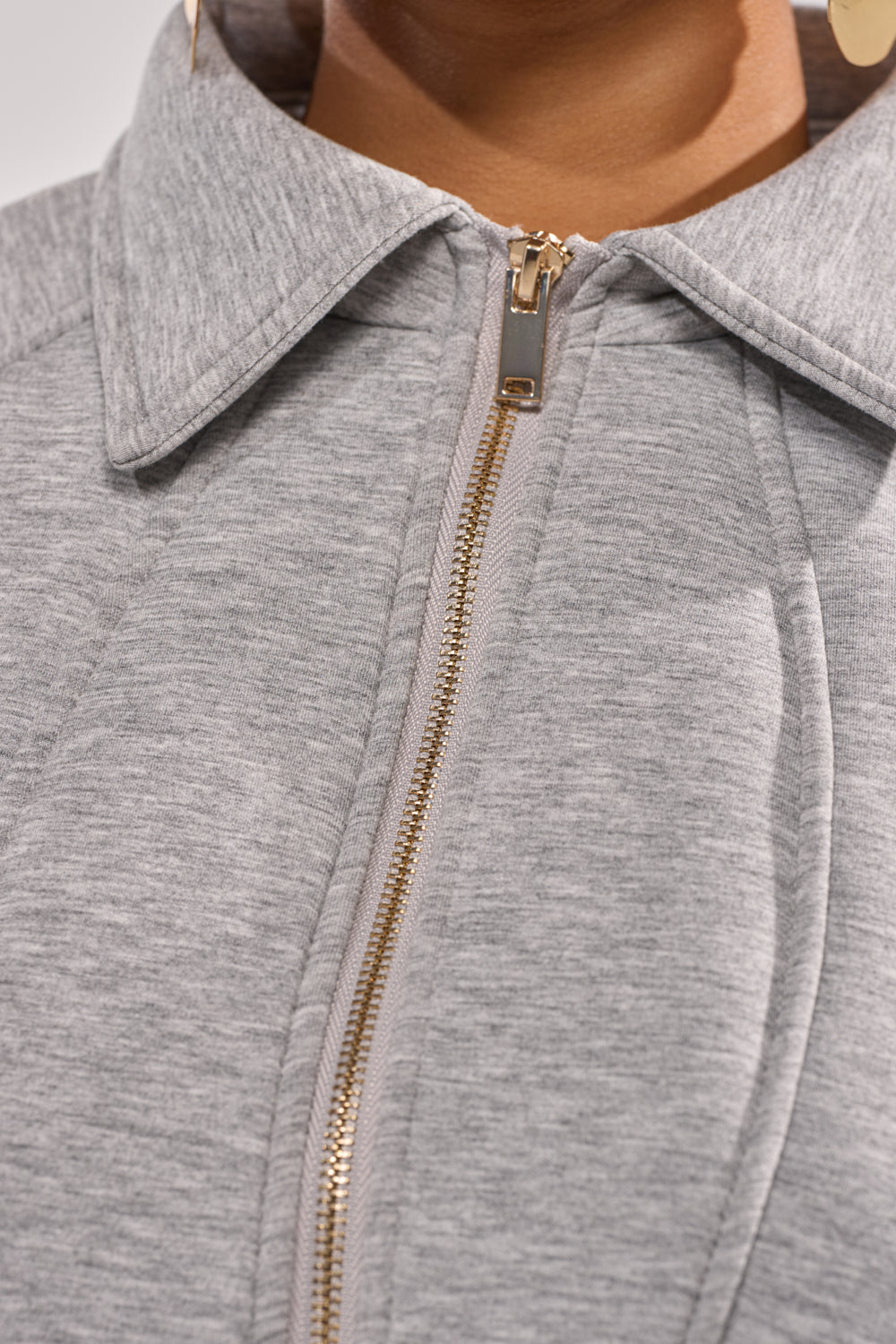Close-up of a person wearing the MONEY TALKS NEOPRENE JACKET, a light gray stretch-knit zip-up with a gold zipper and pointed collar. The soft, textured fabric is highlighted, along with part of a gold earring and the person's neck. 7
