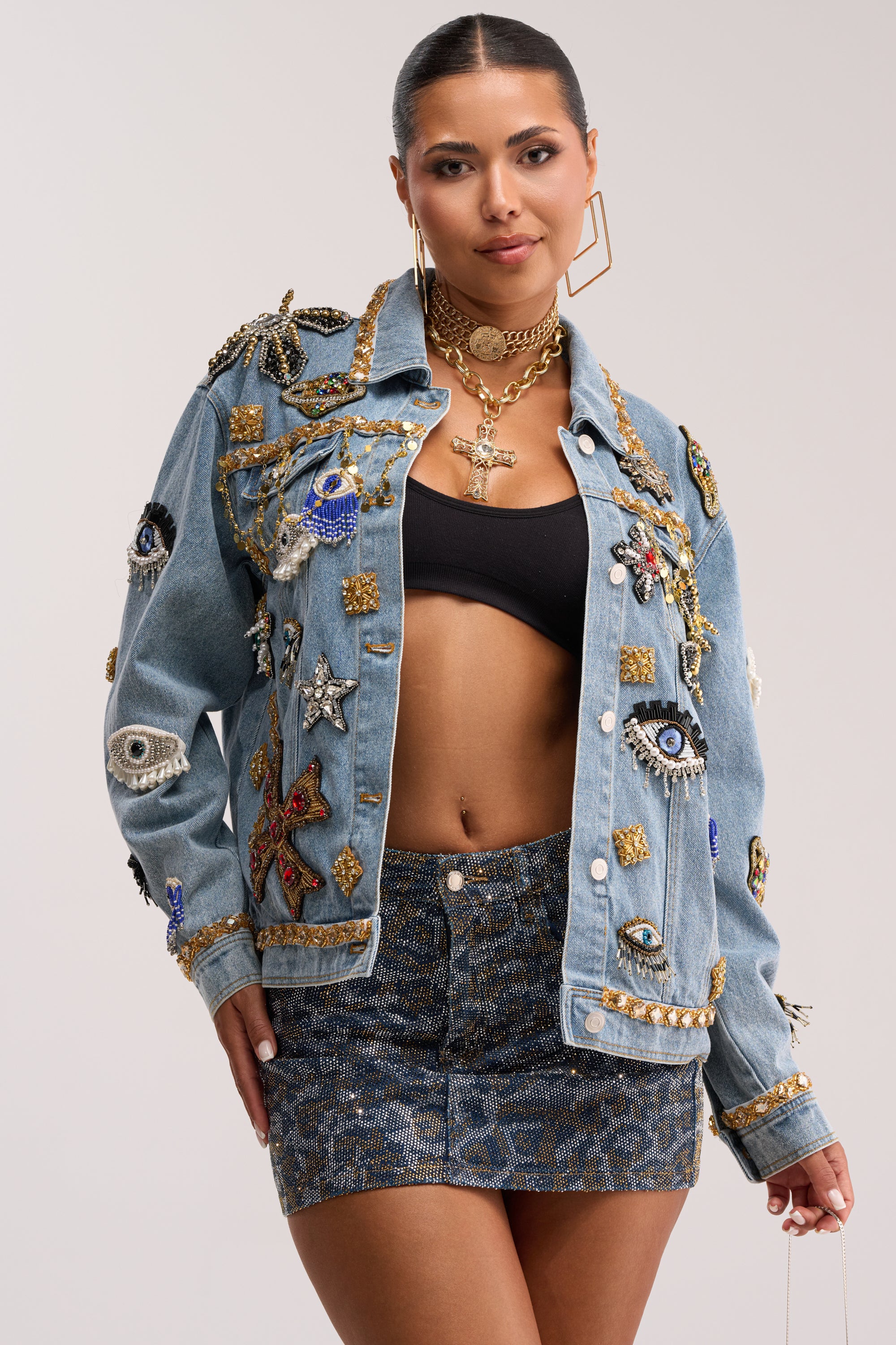 A woman stands confidently in the IN PLAIN SIGHT EMBELLISHED DENIM JACKET adorned with embroidered beading, eyes, stars, and gold accents, paired with a black crop top and a patterned mini skirt.