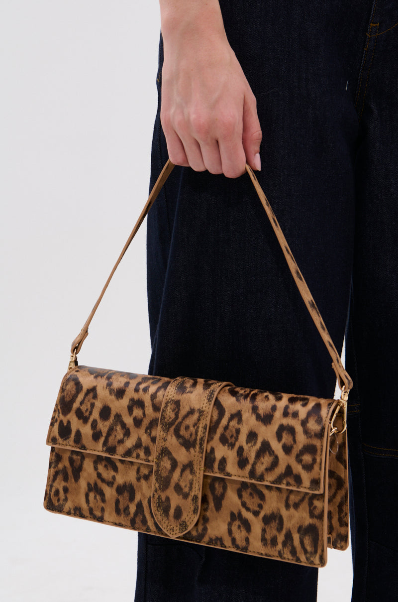 A person in dark pants holds the CAN'T BE TAMED LEOPARD PRINT PURSE—a chic, rectangular leopard print bag with a short strap—against a plain light background.