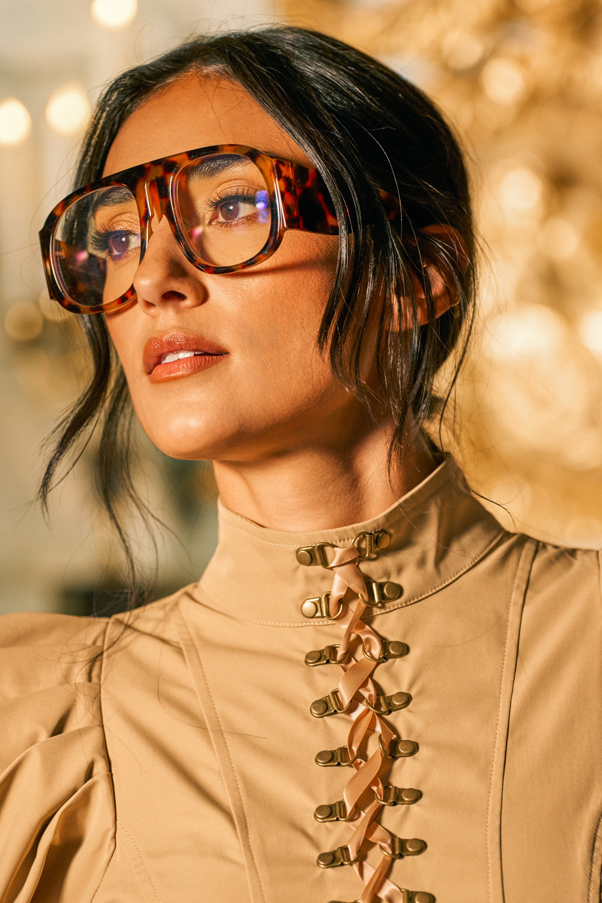 A woman with dark hair wears the SHE MEANS BUSINESS GLASSES—oversized brown tortoise shell frames—paired with a tan high-neck top featuring gold clasps, gazing to the side in soft, warm lighting set against a blurred golden background.
