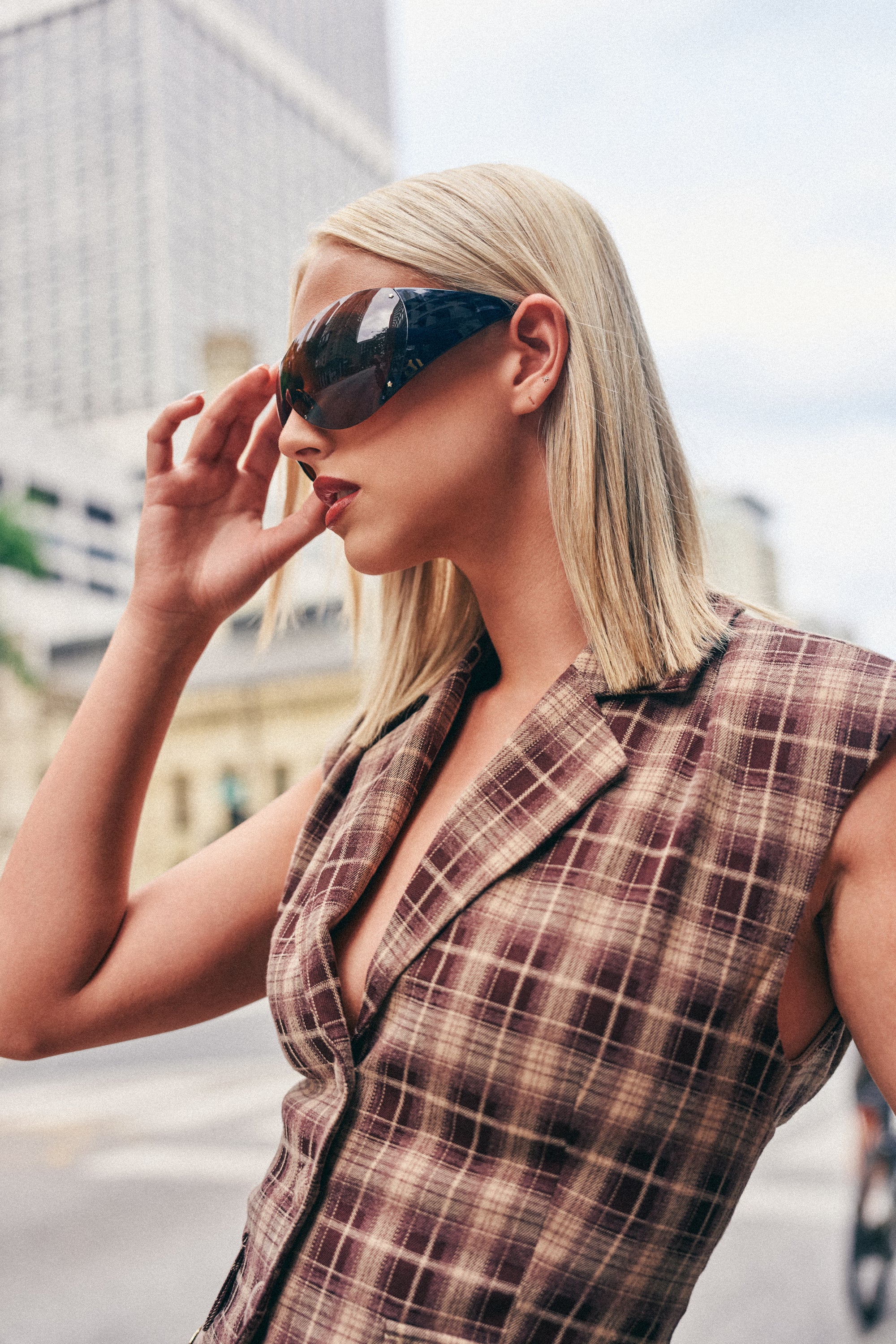A woman with straight blonde hair wears the OUT OF YOUR LEAGUE SUNGLASSES and a sleeveless plaid blazer, standing outdoors in an urban setting with tall buildings in the background.