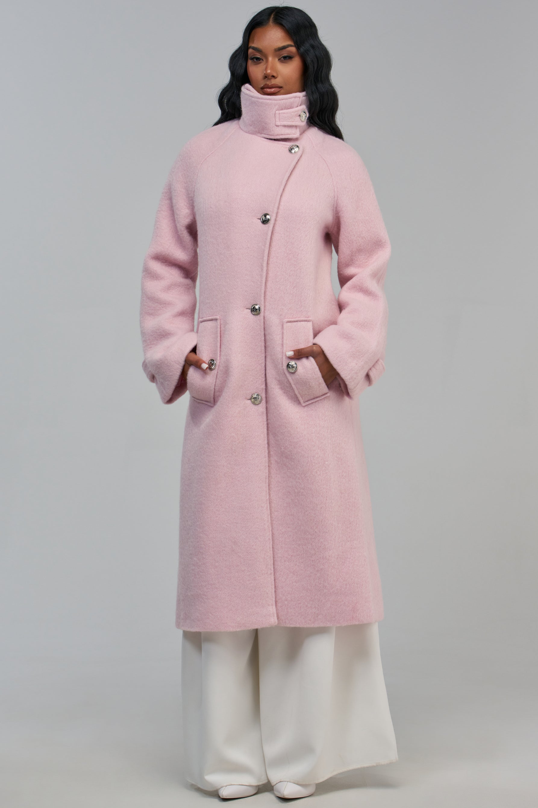 A person wears a longline light pink POPPY WOOL BLEND COAT with a high collar, silver buttons, and pockets, paired with wide-leg white trousers.