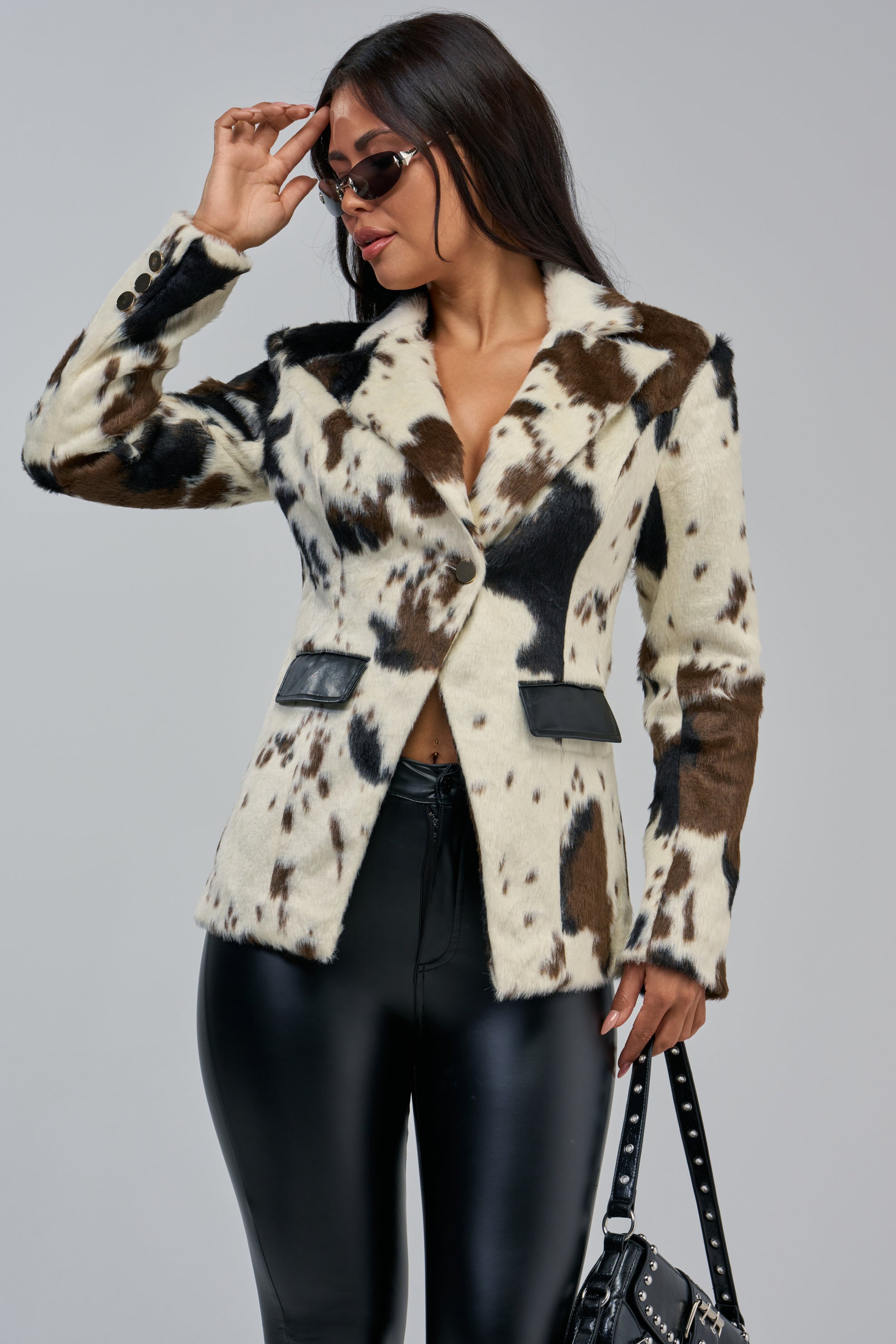 A woman wearing the DIXIE FAUX FUR BLAZER, styled with black leather pants and sunglasses, holds a black studded handbag against a plain light gray background.