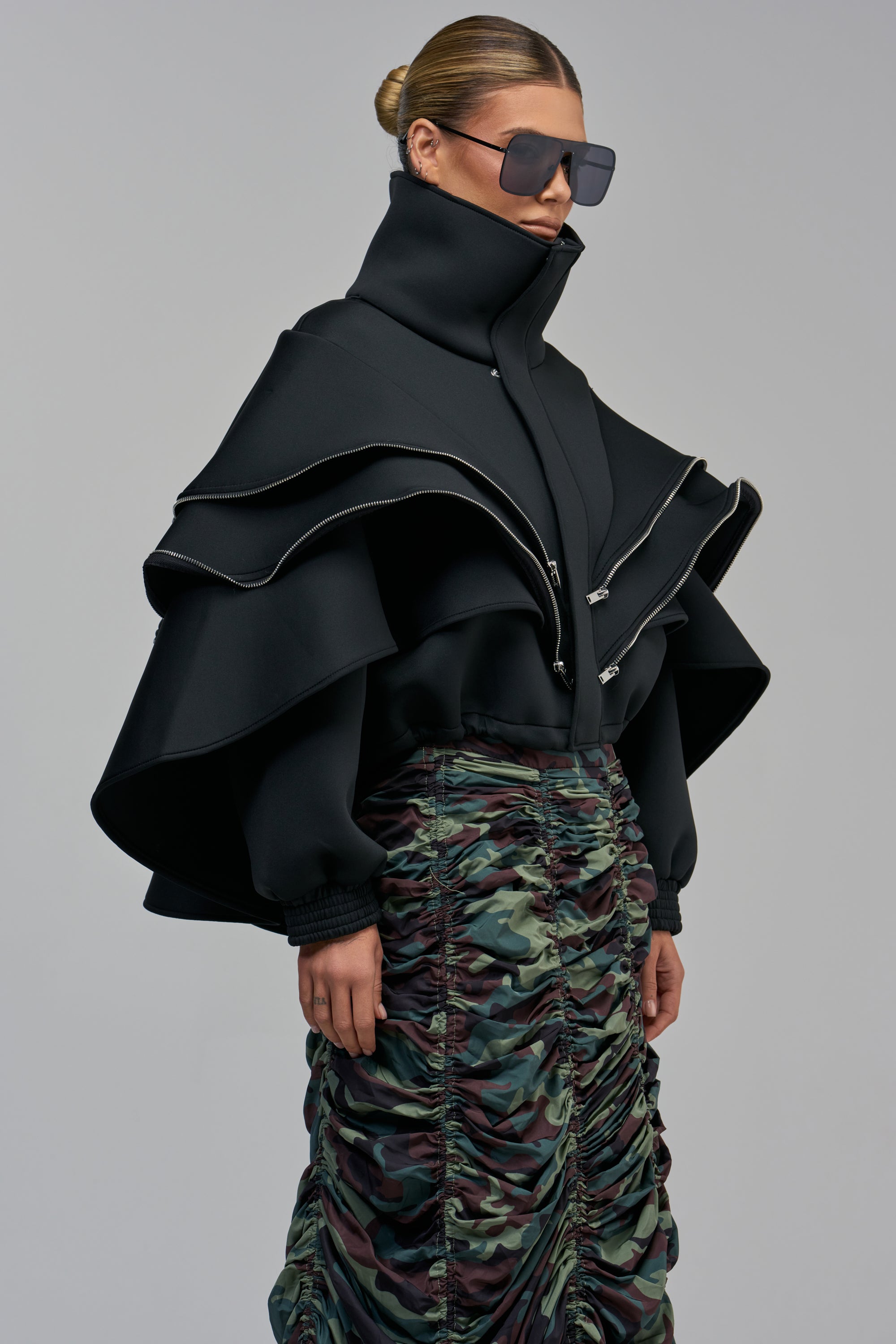 A person wearing a WORK OF ART LAYERED BOMBER IN BLACK with prominent silver zippers and a high collar, paired with an avant-garde layered, camouflage-print skirt.