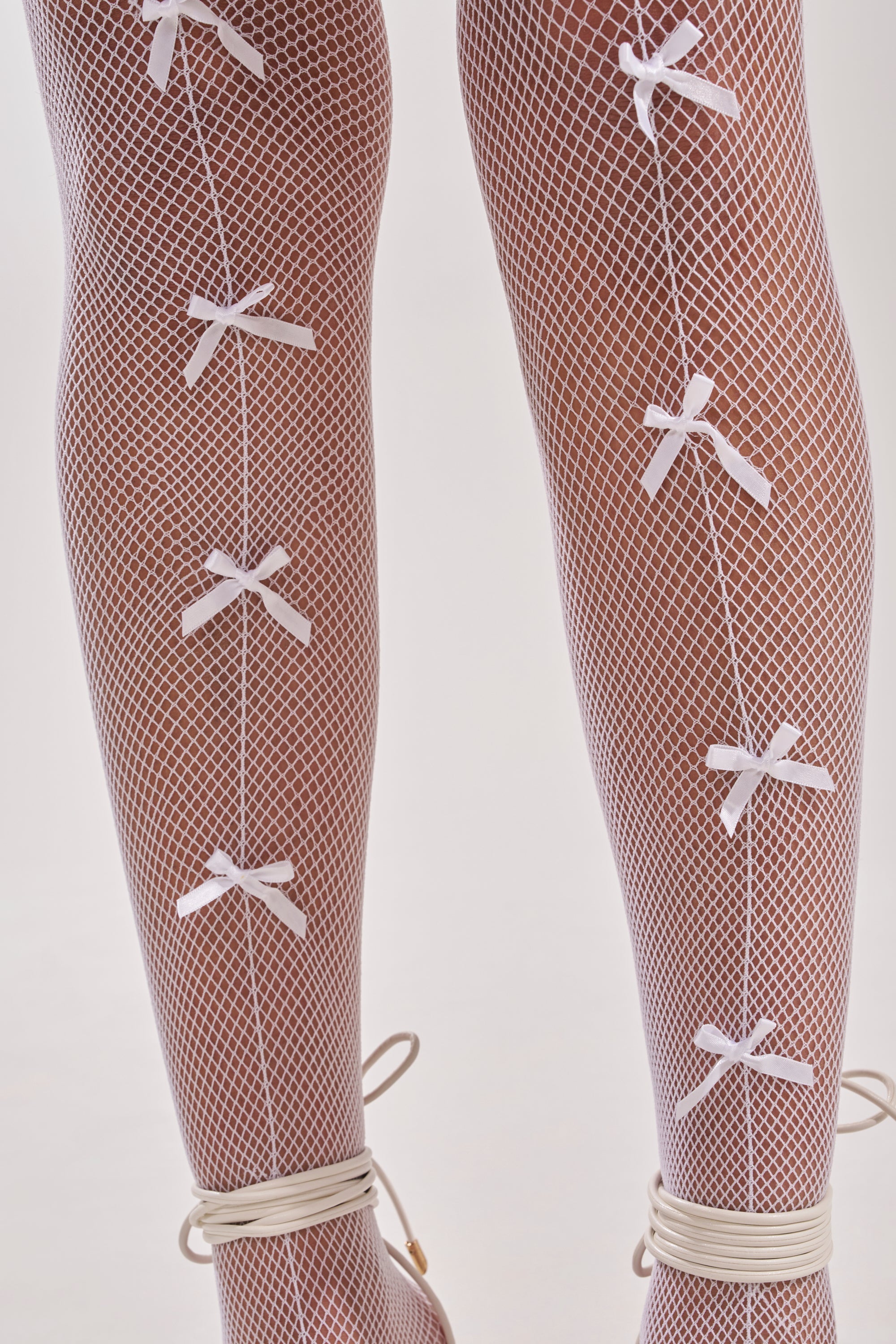 Close-up of legs in WITH A BOW TIGHTS: white fishnet tights featuring vertical rows of small white satin bows and lace-up ankle details. Set against a plain, light background. 2